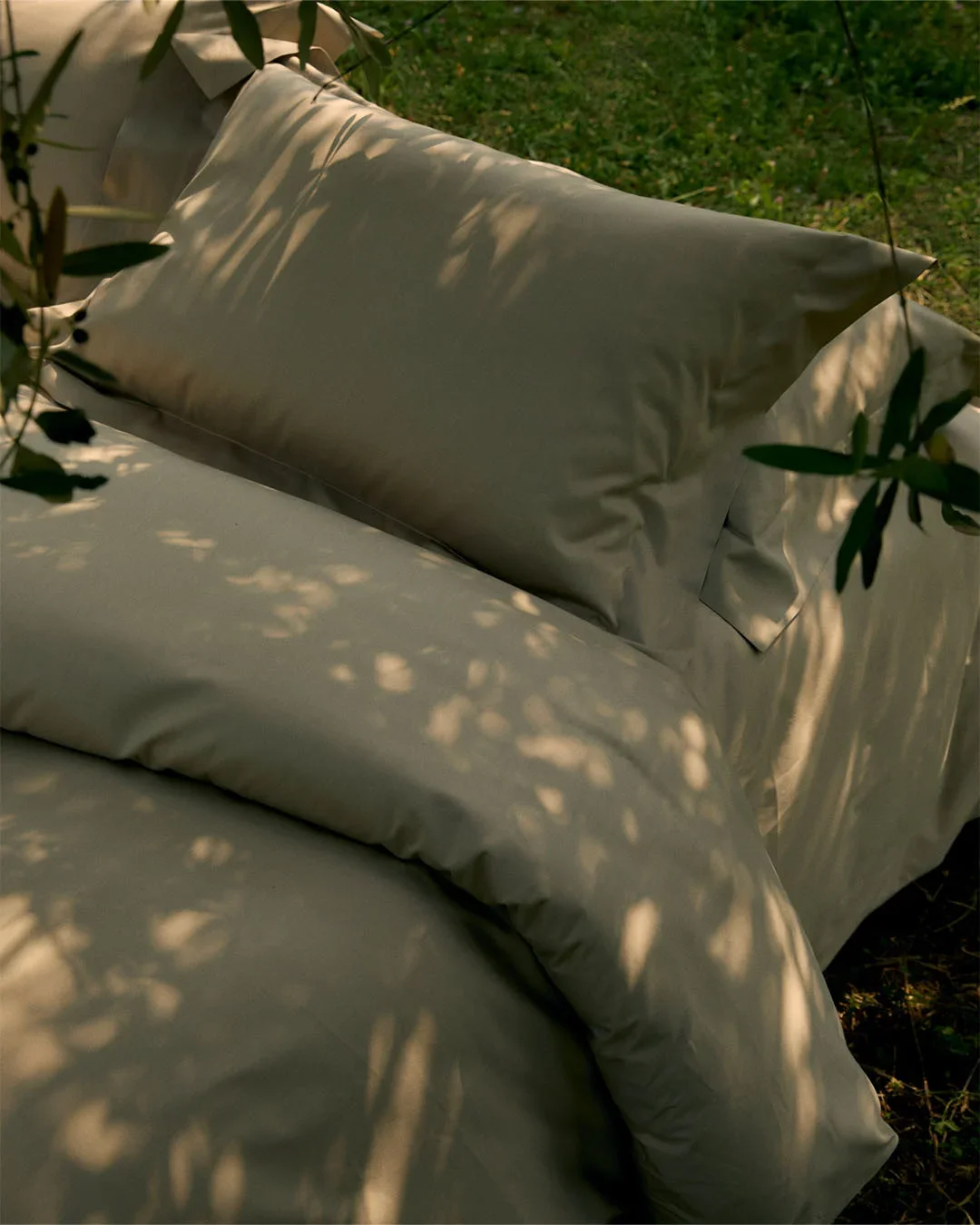 Scenografia outdoor per un set da letto in cotone verde oliva adagiato sul prato, avvolto dai giochi di ombre e dalla luce naturale filtrata dalle fronde di un ulivo