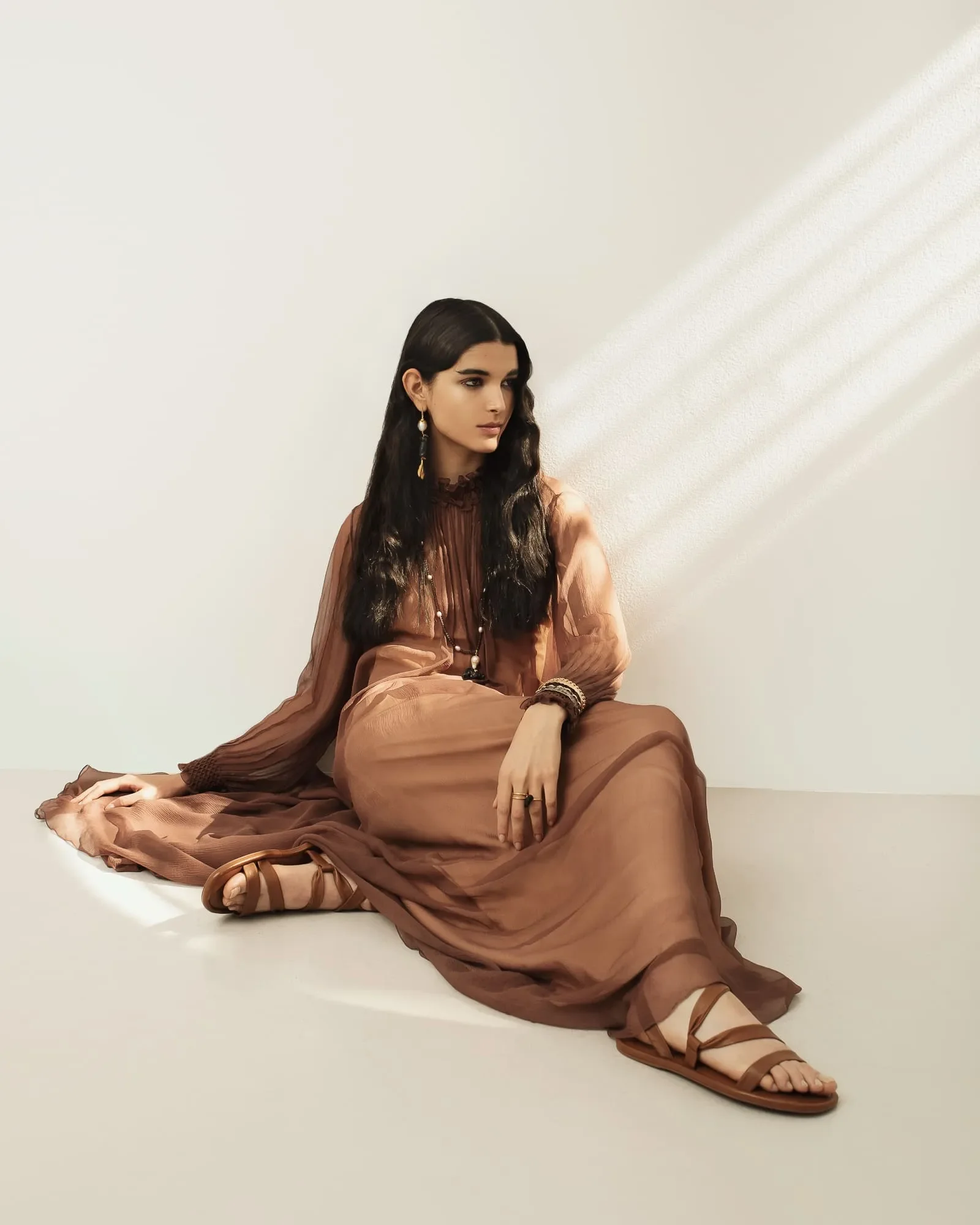 Modella seduta a terra con fluido abito lungo in chiffon marrone cioccolato, sandali in pelle e giochi di luce naturale.