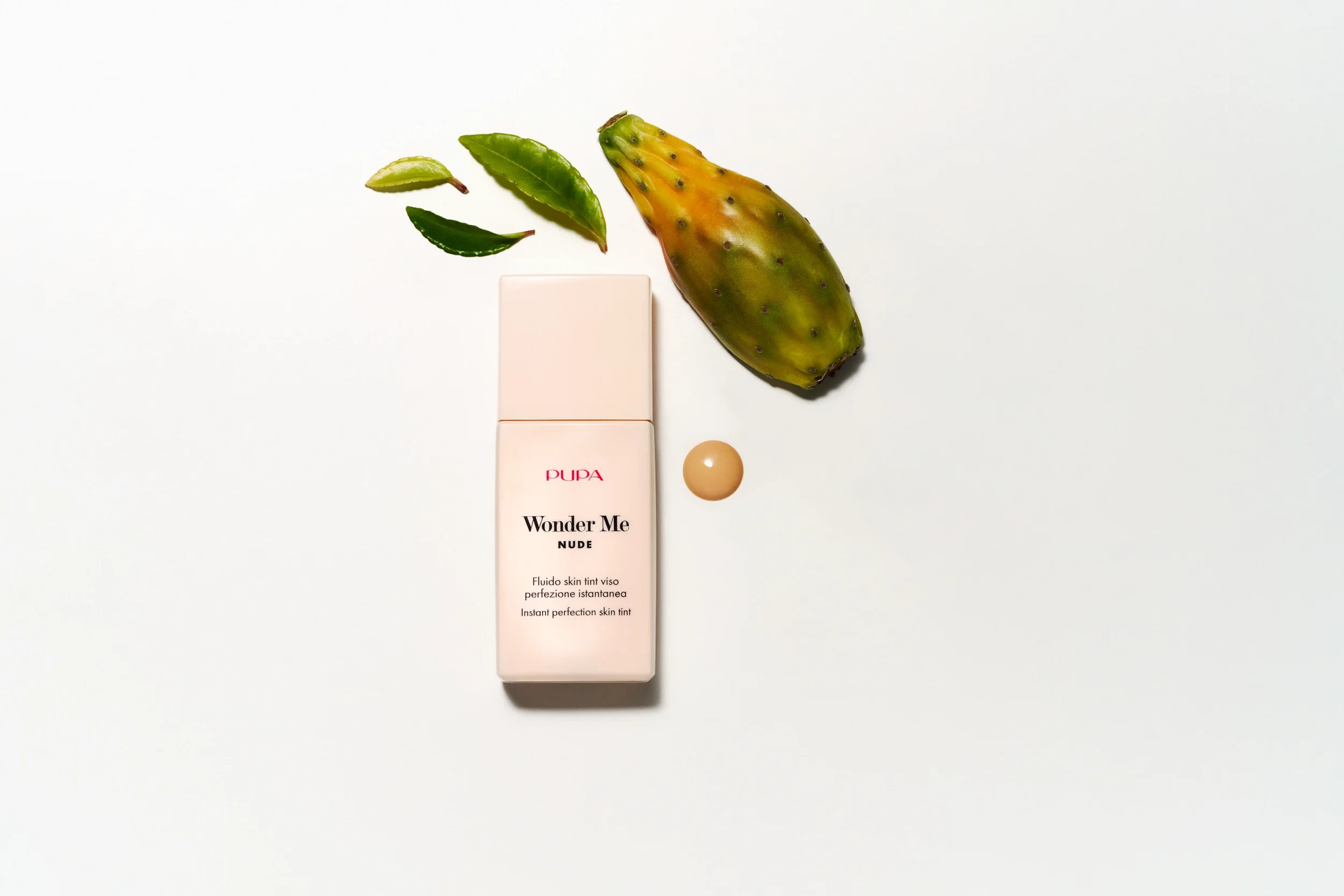 Composizione still life flatlay per la campagna Pupa Wonder Me Nude Skin Tint con flacone di prodotto, singola goccia di fluido, fico d'India e foglie verdi fresche su sfondo bianco.