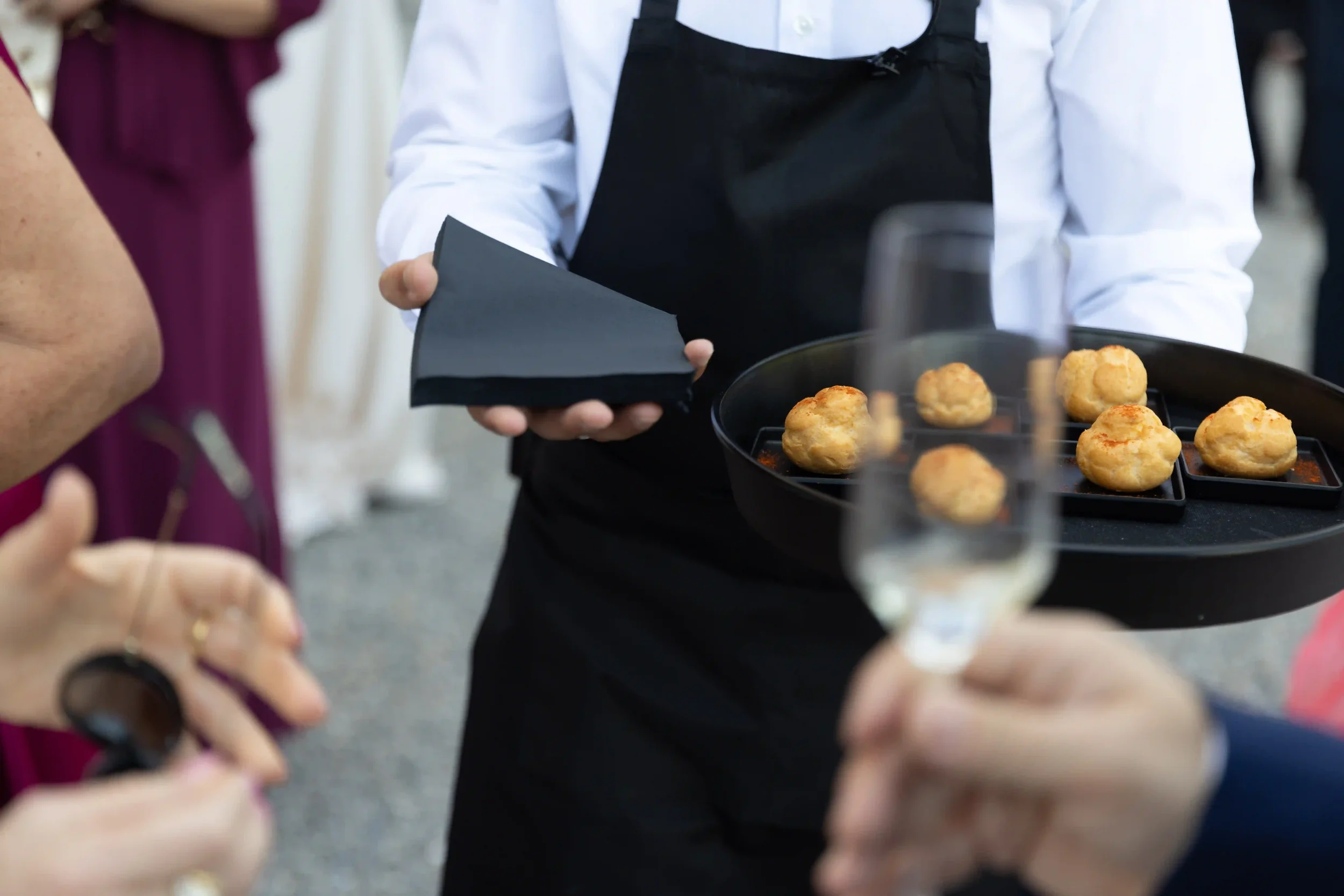 Dettaglio del servizio catering durante l'aperitivo del matrimonio, con cameriere in grembiule nero che serve finger food raffinati e choux salati su vassoio elegante.