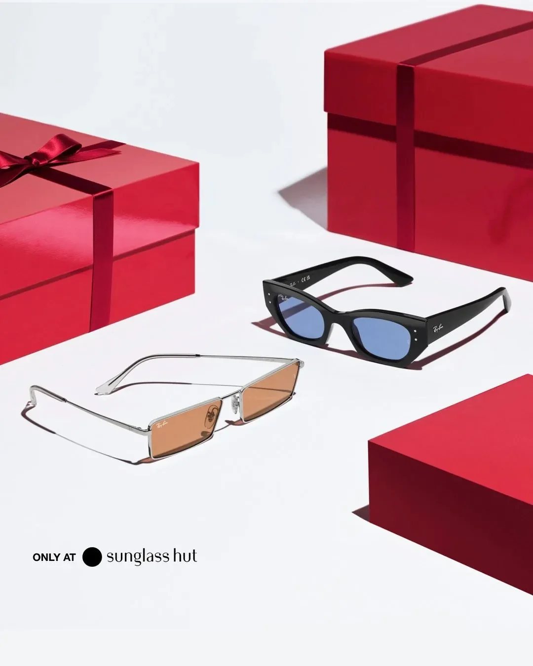 Composizione still life per la campagna Sunglass Hut con due paia di occhiali da sole Ray-Ban, uno in metallo argento con lenti arancioni e uno nero con lenti azzurre, disposti su sfondo bianco tra pacchi regalo rossi natalizi.