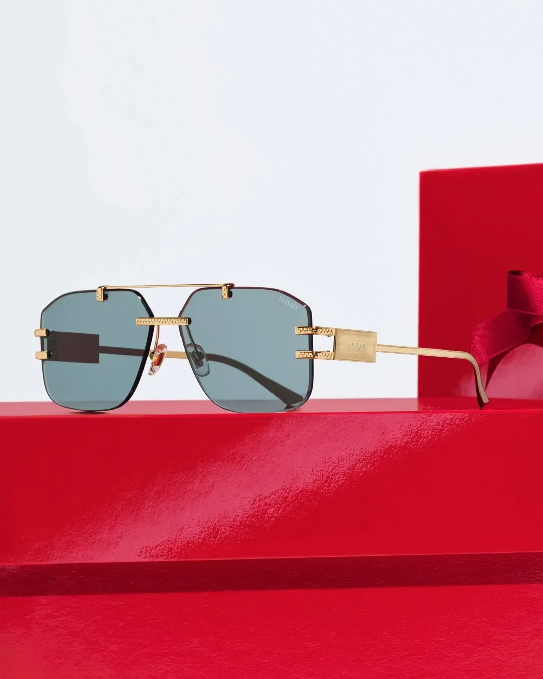 Fotografia still life di occhiali da sole Versace con montatura geometrica in metallo dorato e lenti azzurre, esposti su un gradino di presentazione rosso per la campagna Sunglass Hut.