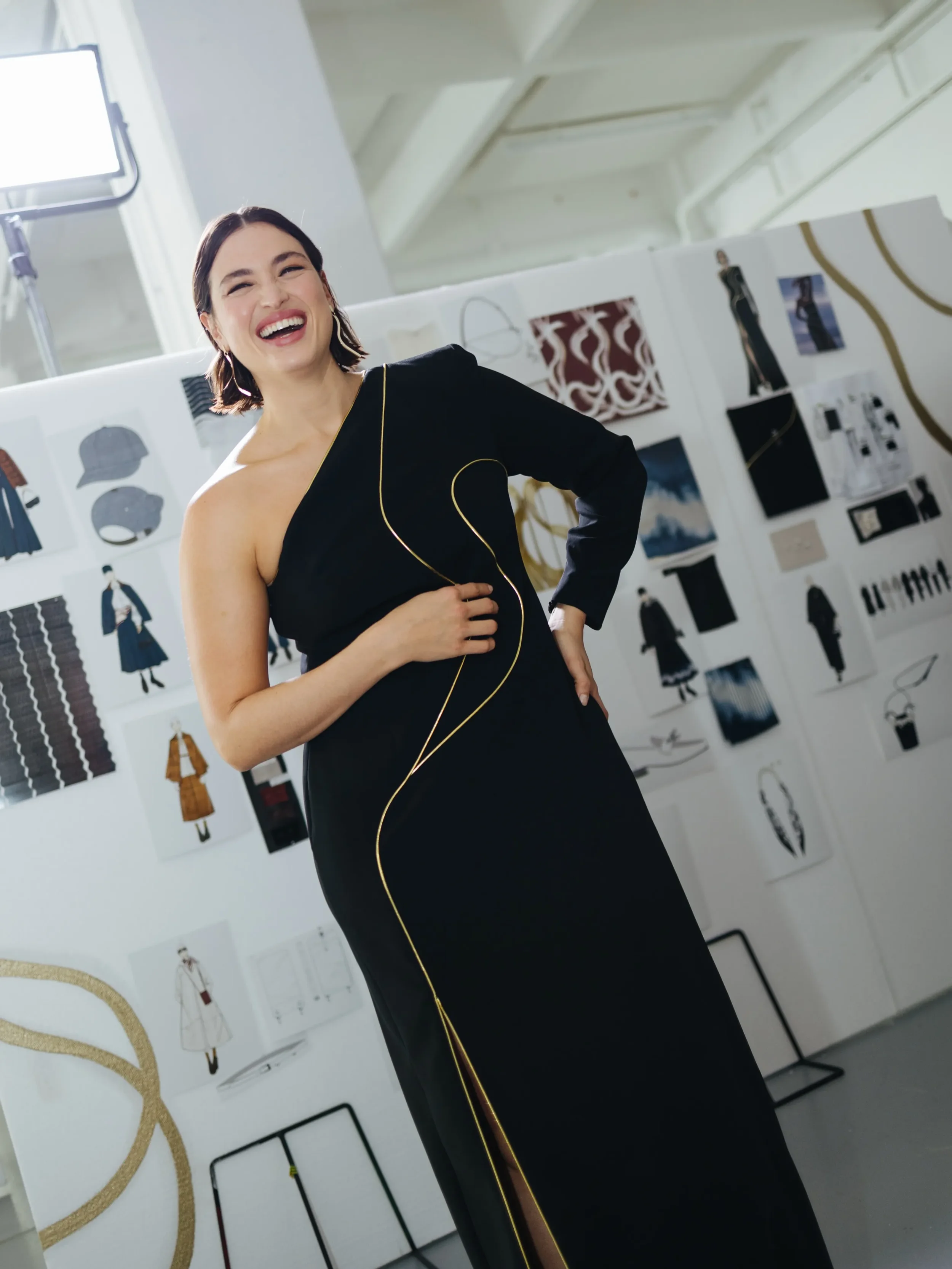 Modella sorridente posa nel backstage della sfilata Marina Rinaldi indossando un abito lungo nero monospalla con profili dorati, davanti al pannello allestito con bozzetti di collezione e moodboard