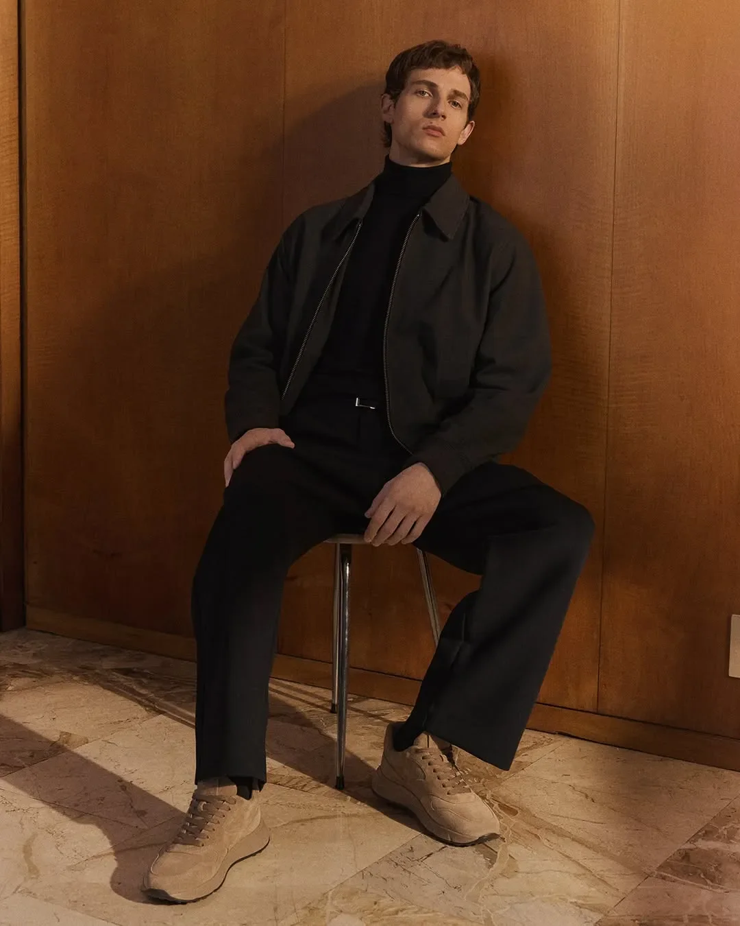 Set design e scenografia per scatto moda maschile Hogan. Modello uomo seduto su sgabello vintage con look scuro e sneakers in camoscio beige