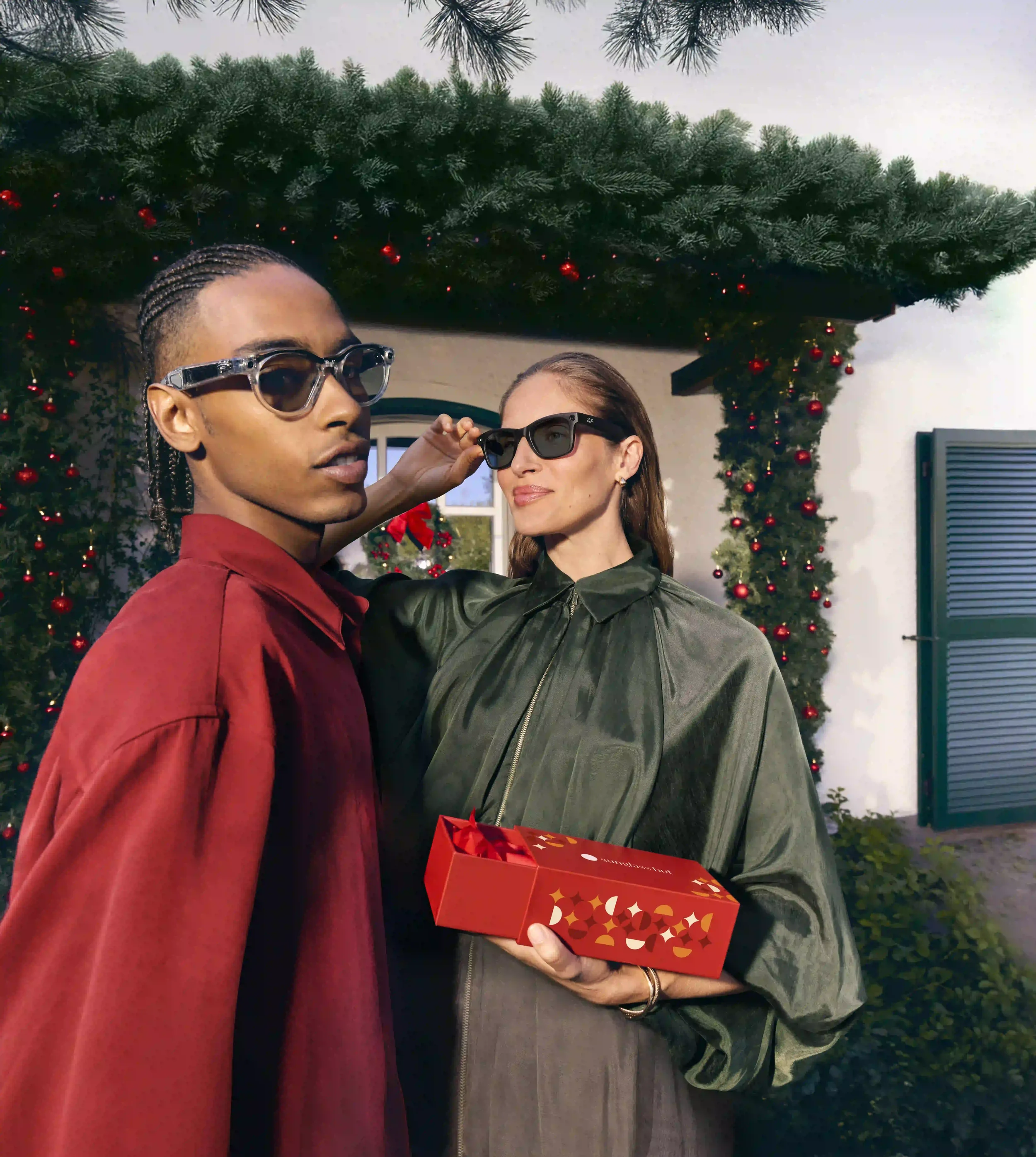 Ritratto promozionale natalizio Sunglass Hut con coppia in posa sotto una ghirlanda di pino decorata con palline rosse, con scatola regalo e occhiali da sole moderni con montatura trasparente e nera.