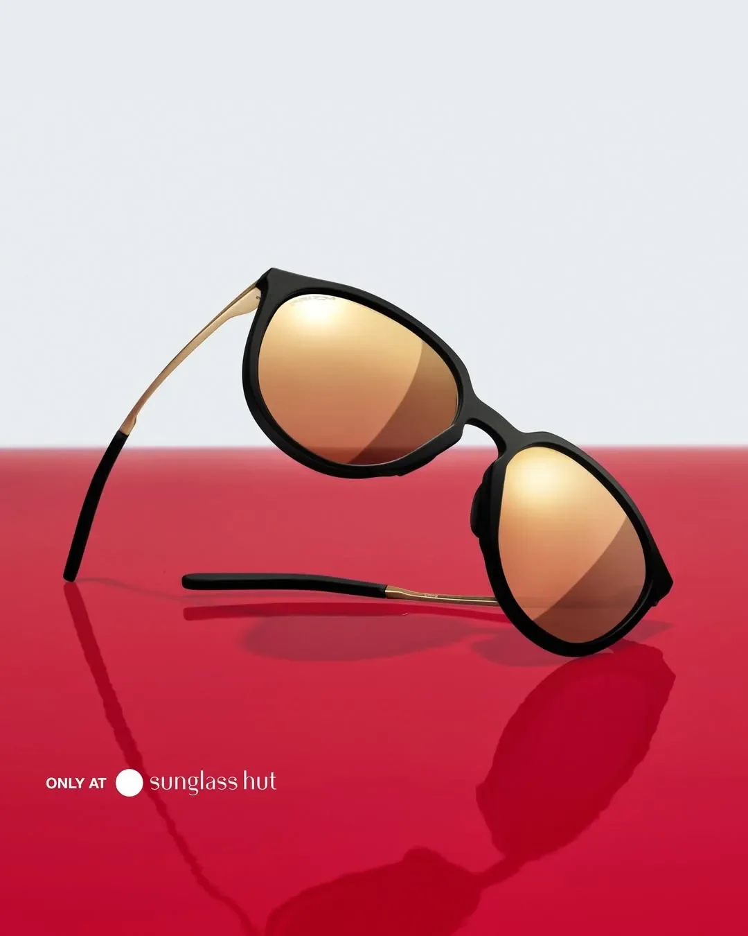 Fotografia still life di occhiali da sole Oakley con montatura nera opaca e lenti specchiate oro Prizm, adagiati su una superficie rossa brillante con riflesso grafico per Sunglass Hut.