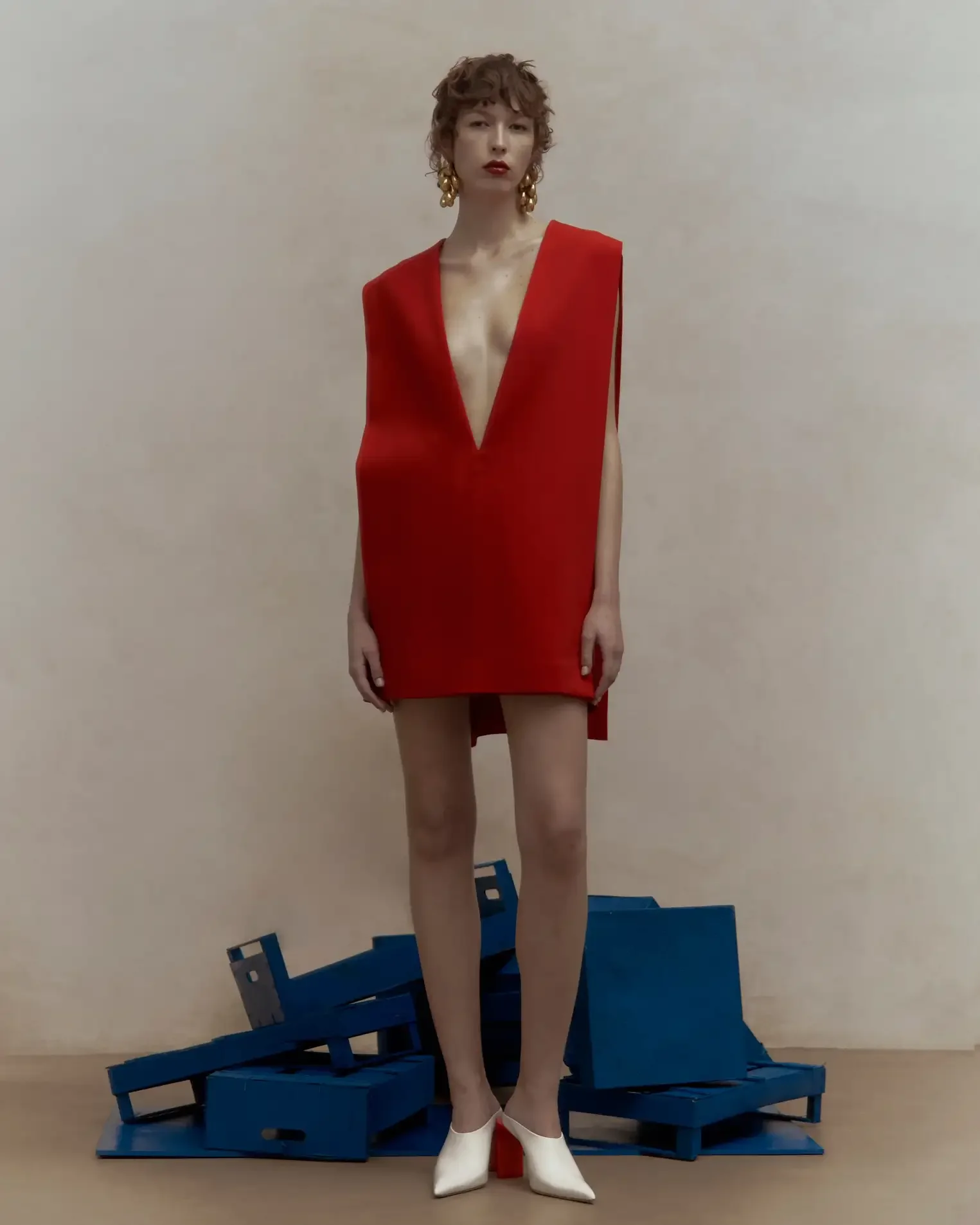 Modella con abito scultoreo rosso acceso dal profondo scollo a V, orecchini dorati e sabot bianchi con tacco sagomato rosso, in piedi sopra una pila di cassette in legno blu su sfondo neutro. Styling editoriale per Le Mile Magazine.