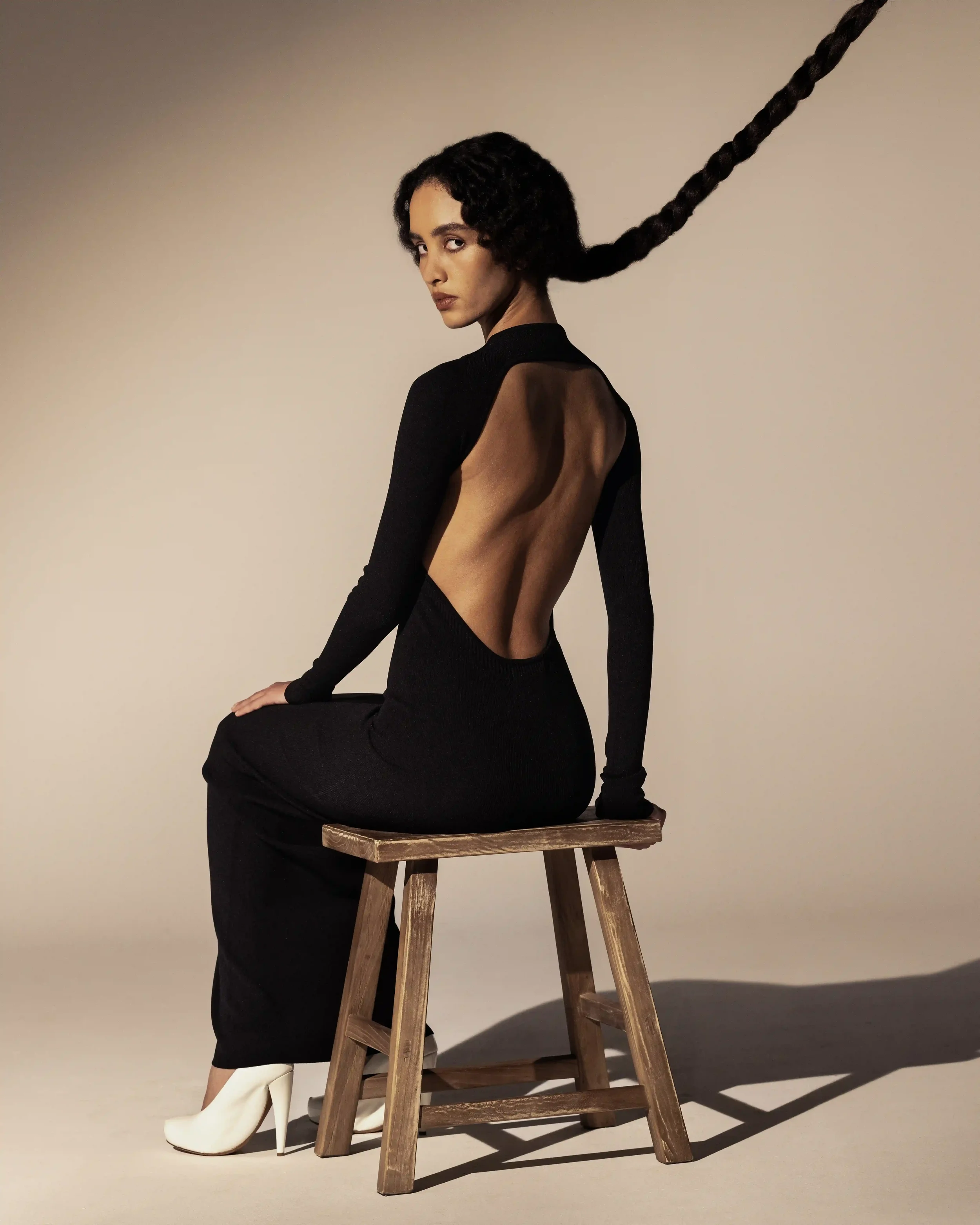 Set design essenziale con sgabello in legno grezzo per lo shooting Laneus. Modella seduta con abito lungo nero scollato sulla schiena, décolleté bianche e treccia sollevata.