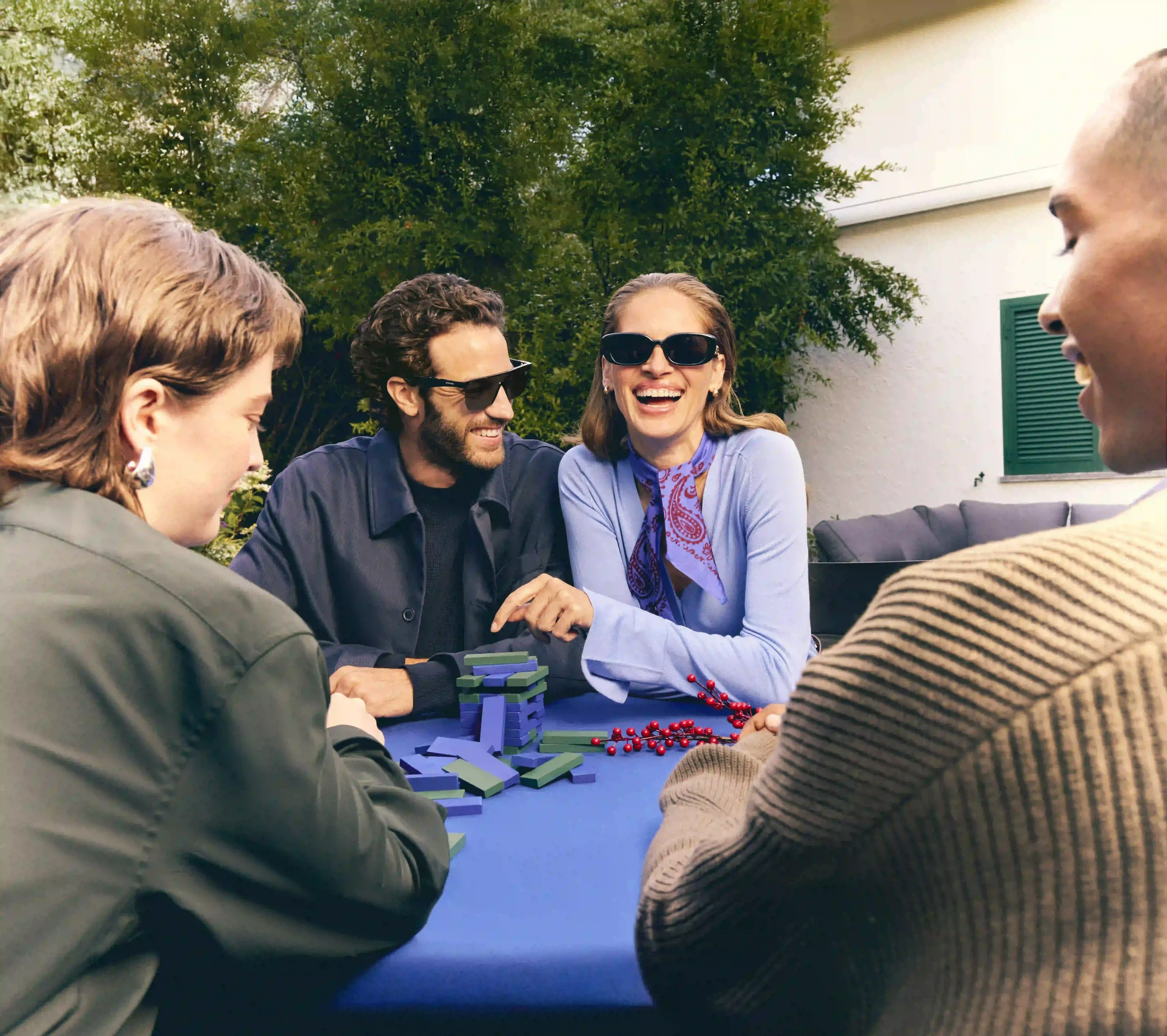 Scatto lifestyle e di festa per Sunglass Hut con gruppo di amici sorridenti che giocano a Jenga all'aperto attorno a un tavolo blu, indossando occhiali da sole da uomo e donna.