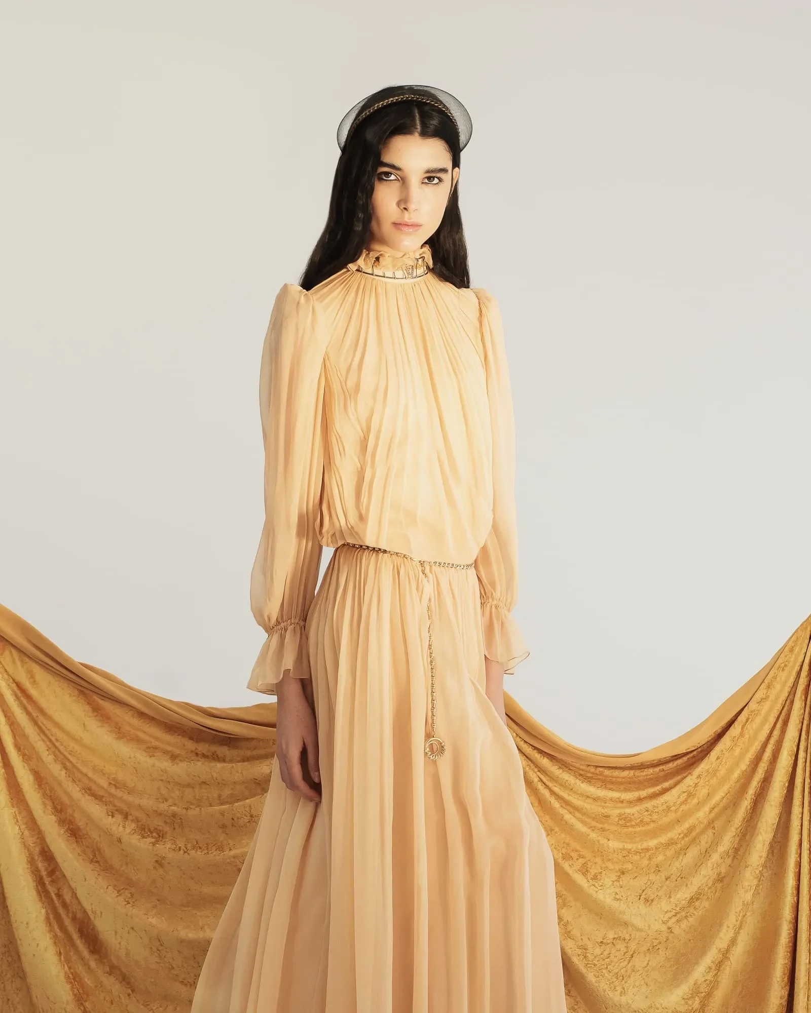 Ritratto a figura intera di una modella in elegante abito lungo plissé color cipria con cintura a catena dorata e drappeggio in velluto giallo.