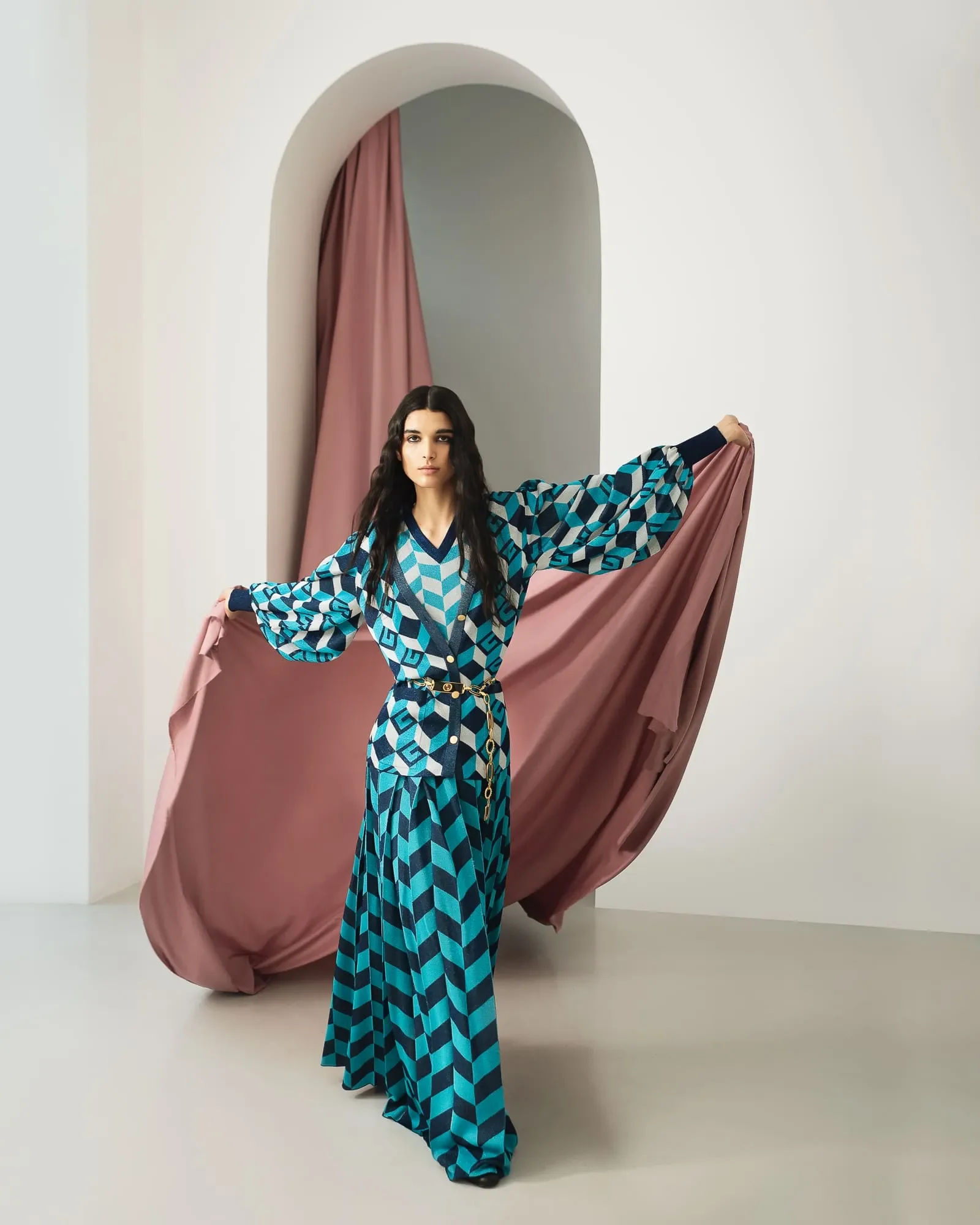 Modella in completo in maglia a motivi geometrici ottanio e blu con cintura dorata, mentre apre un drappeggio rosa antico sotto un arco scenografico.