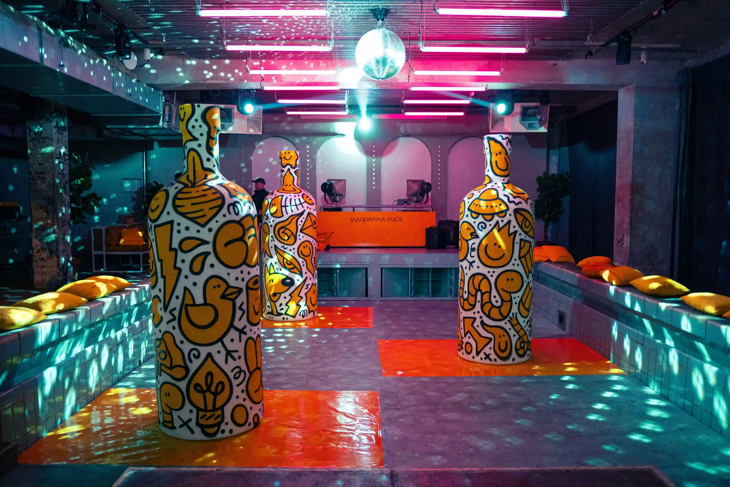 Allestimento scenografico per il party Mandarina Duck a Berlino all'interno di una piscina dismessa con luci al neon rosa, disco ball e grandi sculture pop art.