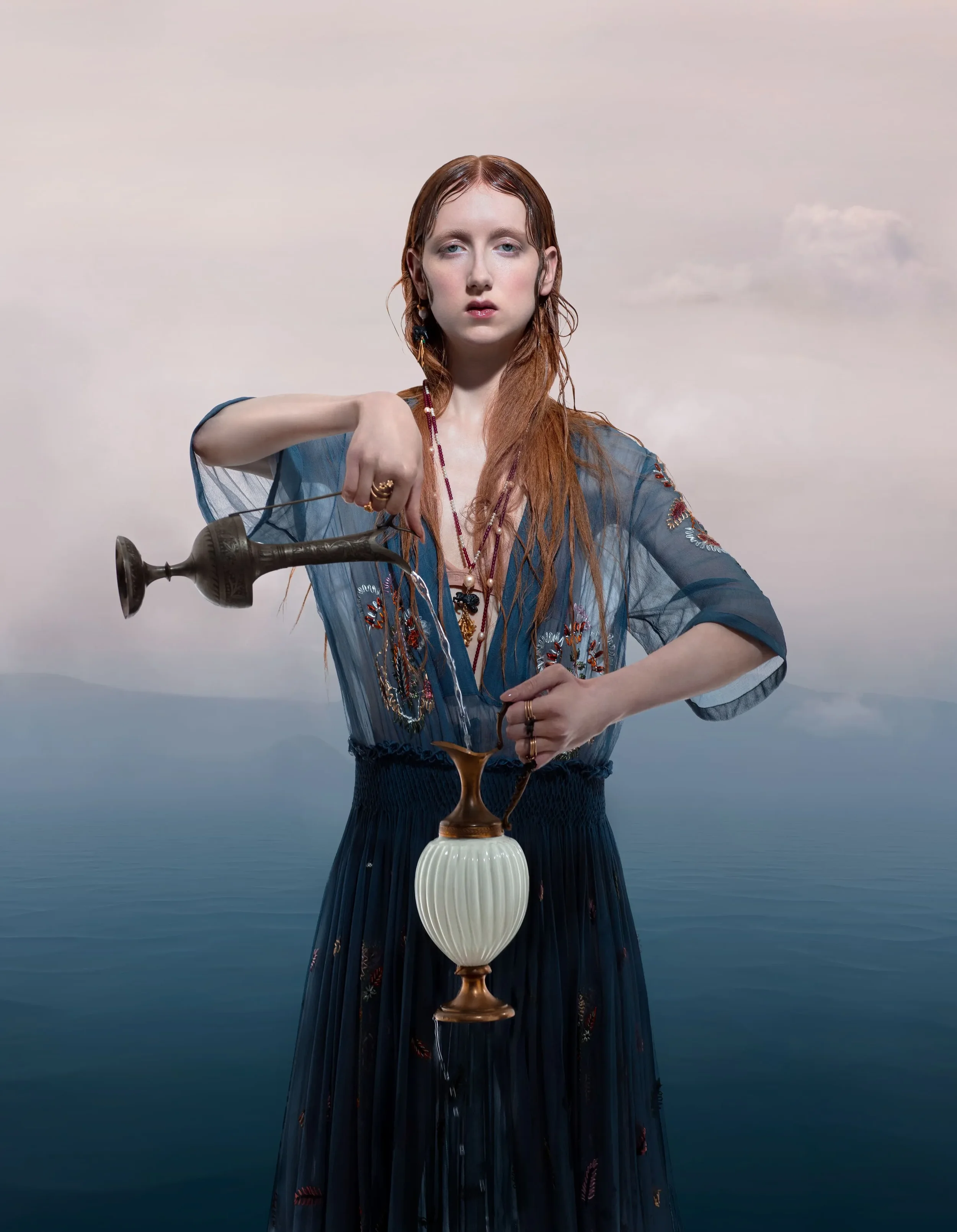 Ritratto editoriale per Vogue Portugal con modella dal wet look in abito blu trasparente ricamato, intenta a versare acqua da un'anfora d'epoca su uno sfondo marino.