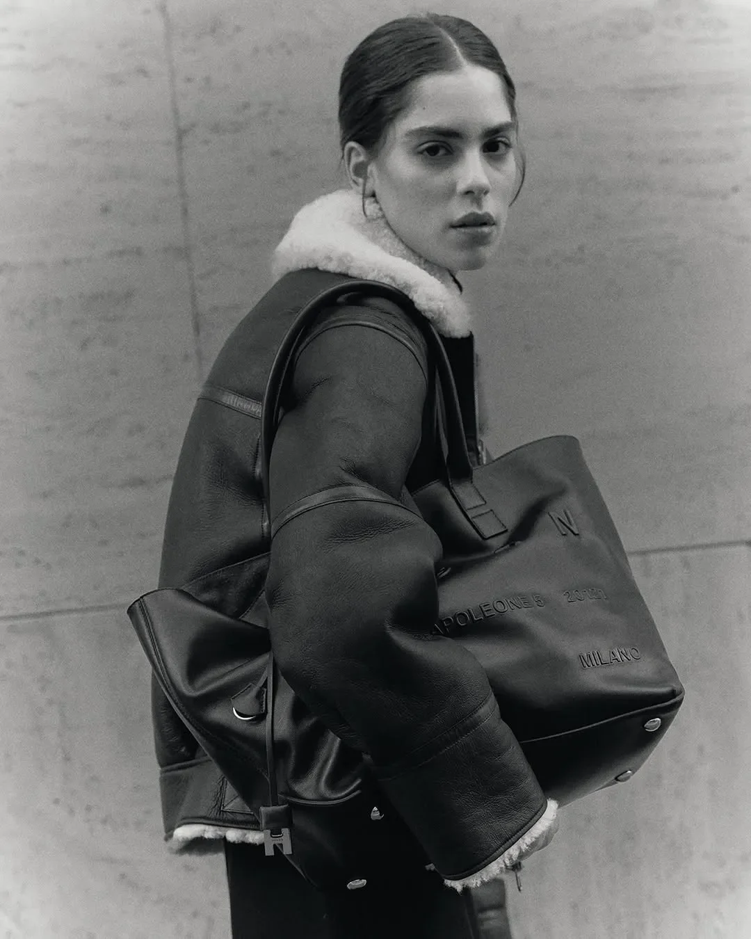 Ritratto di moda in bianco e nero per la campagna ADV Hogan. Modella con giacca in montone e borsa oversize in pelle su sfondo architettonico essenziale.