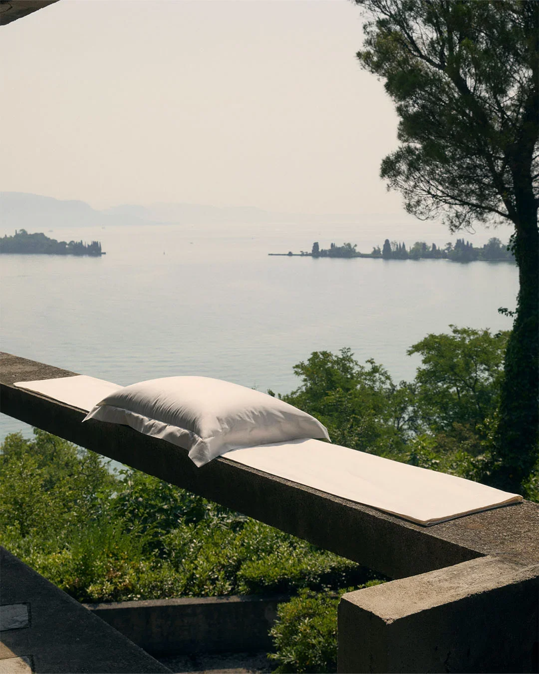 Allestimento scenografico e props styling con cuscino in lino bianco posizionato lungo la balaustra in pietra di una terrazza, con vista panoramica sul lago e colline al tramonto