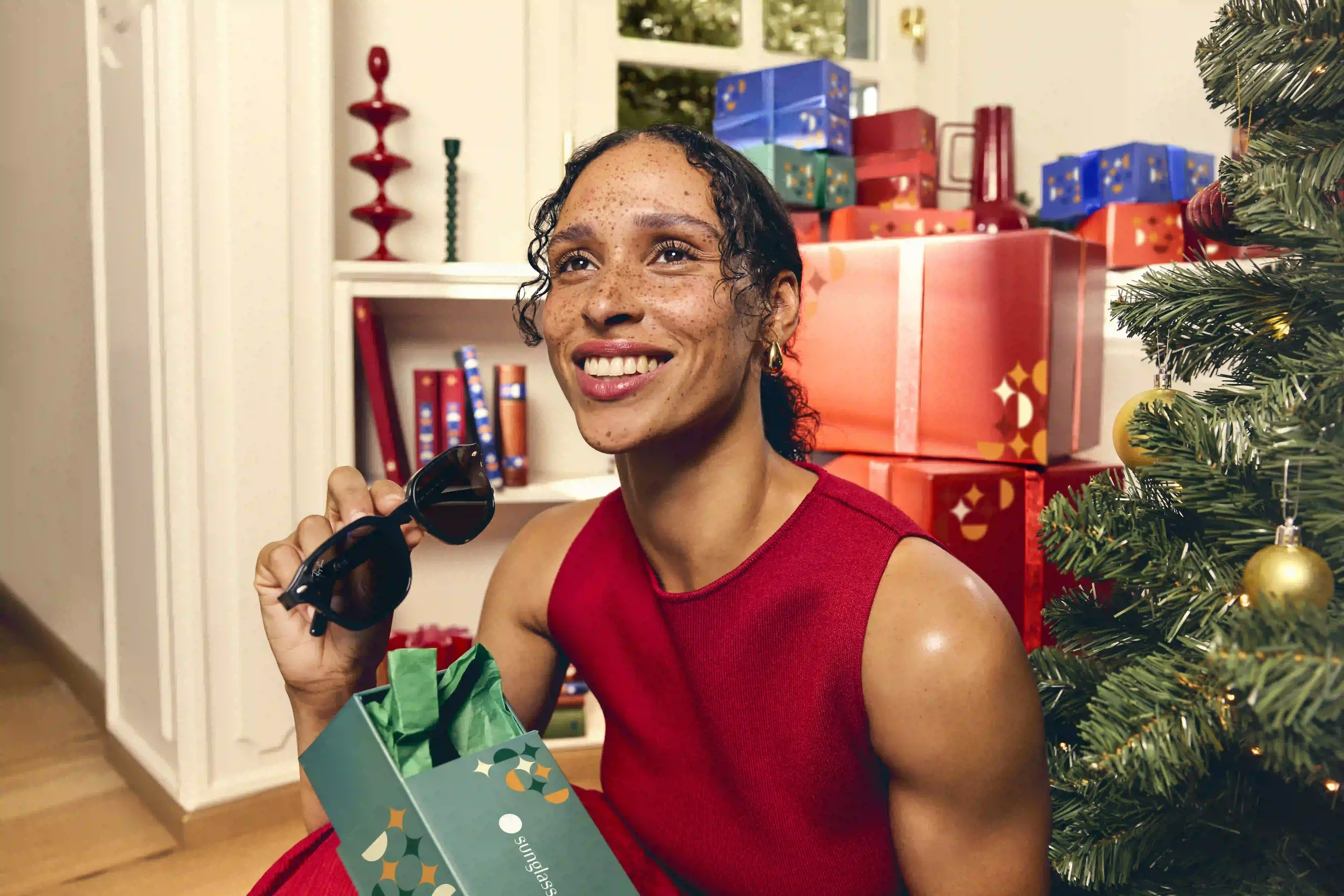 Scatto lifestyle della campagna natalizia Sunglass Hut con modella in abito rosso che scopre un paio di occhiali da sole da una scatola regalo verde accanto all'albero di Natale."