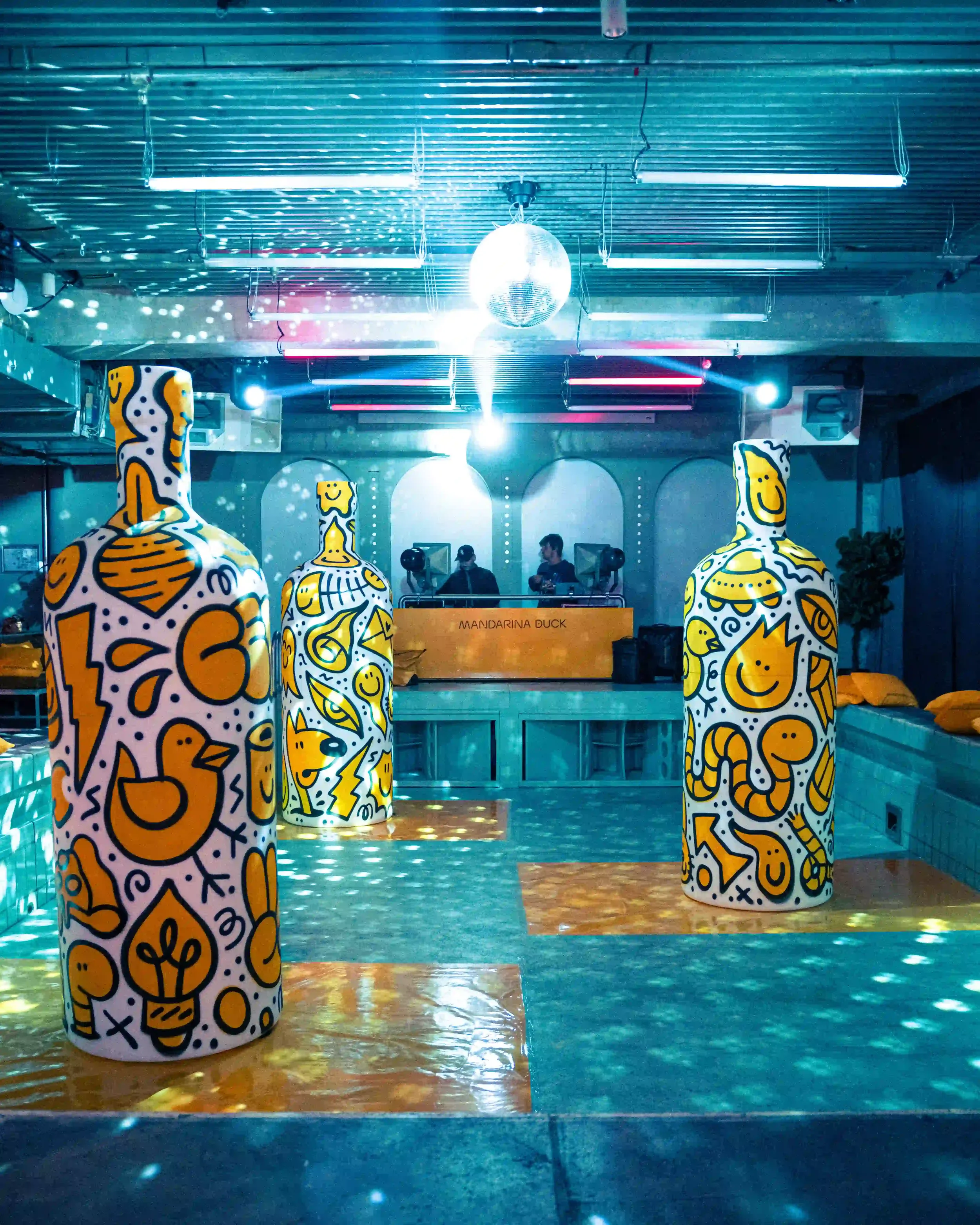 Set design della sala principale per l'evento Mandarina Duck a Berlino, con grandi sculture a forma di bottiglia decorate da graffiti giallo-neri e console DJ con strobosfera