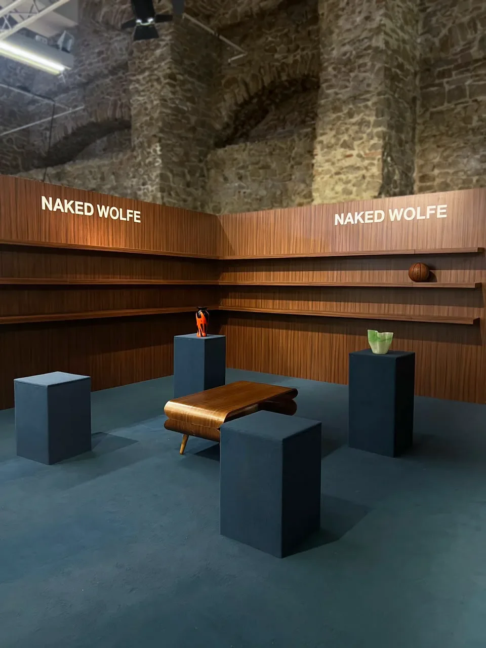 Progetto di space design per il pop-up Naked Wolfe con parete in boiserie di legno scuro e logo illuminato, moquette blu scuro, piedistalli espositivi, tavolino curvo vintage e oggetti scenici in un'architettura storica con volte in pietra.