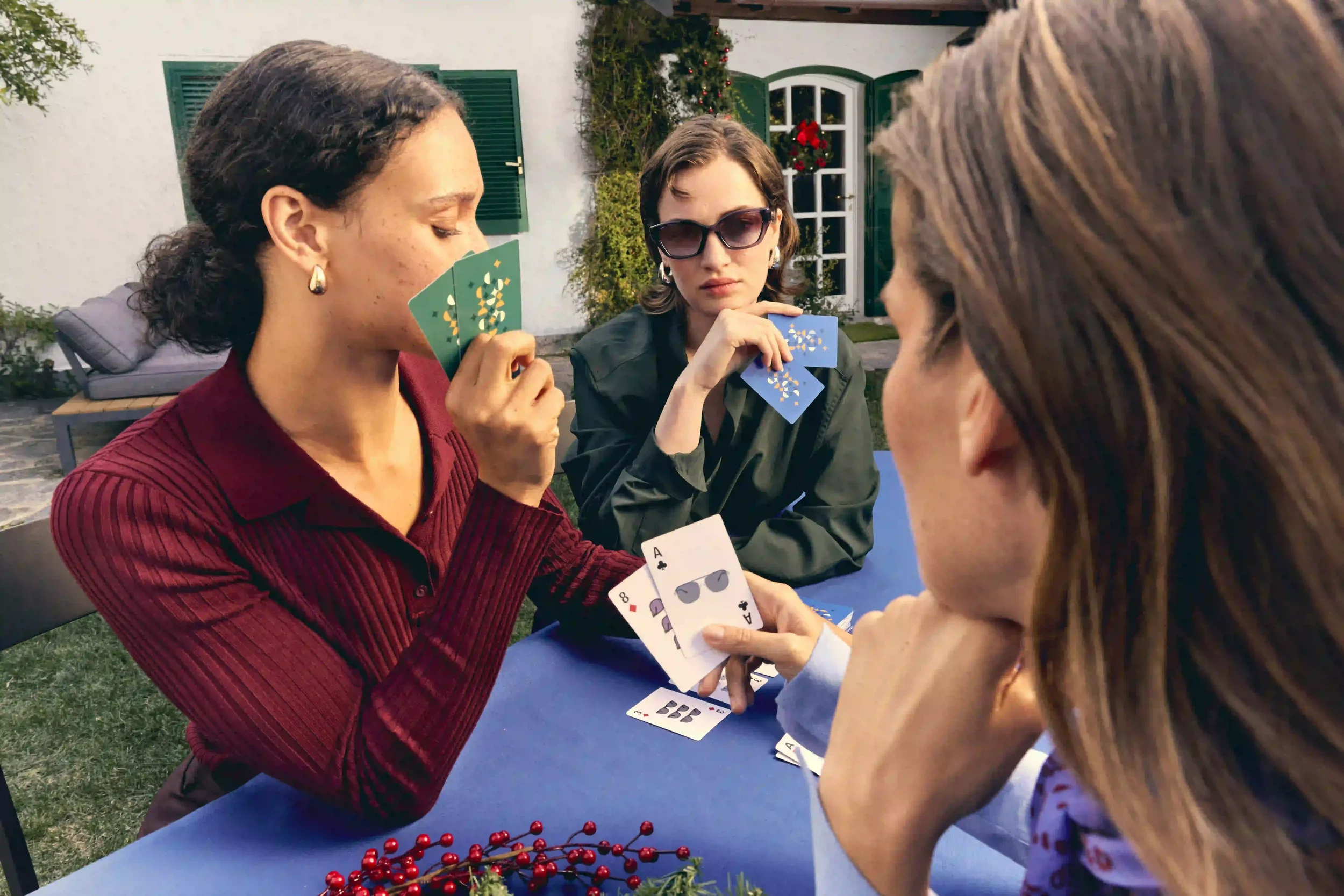 Fotografia di moda e lifestyle per il Natale Sunglass Hut con amiche che giocano a carte in giardino, in un'atmosfera conviviale festiva, indossando occhiali da sole squadrati.