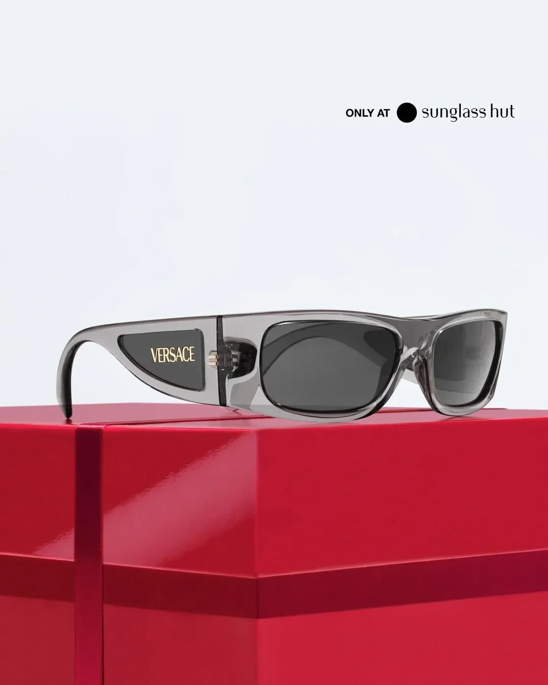 Vista laterale in dettaglio di occhiali da sole Versace in acetato grigio trasparente con logo dorato sull'asta, adagiati su una pacco regalo rosso per la campagna Sunglass Hut.