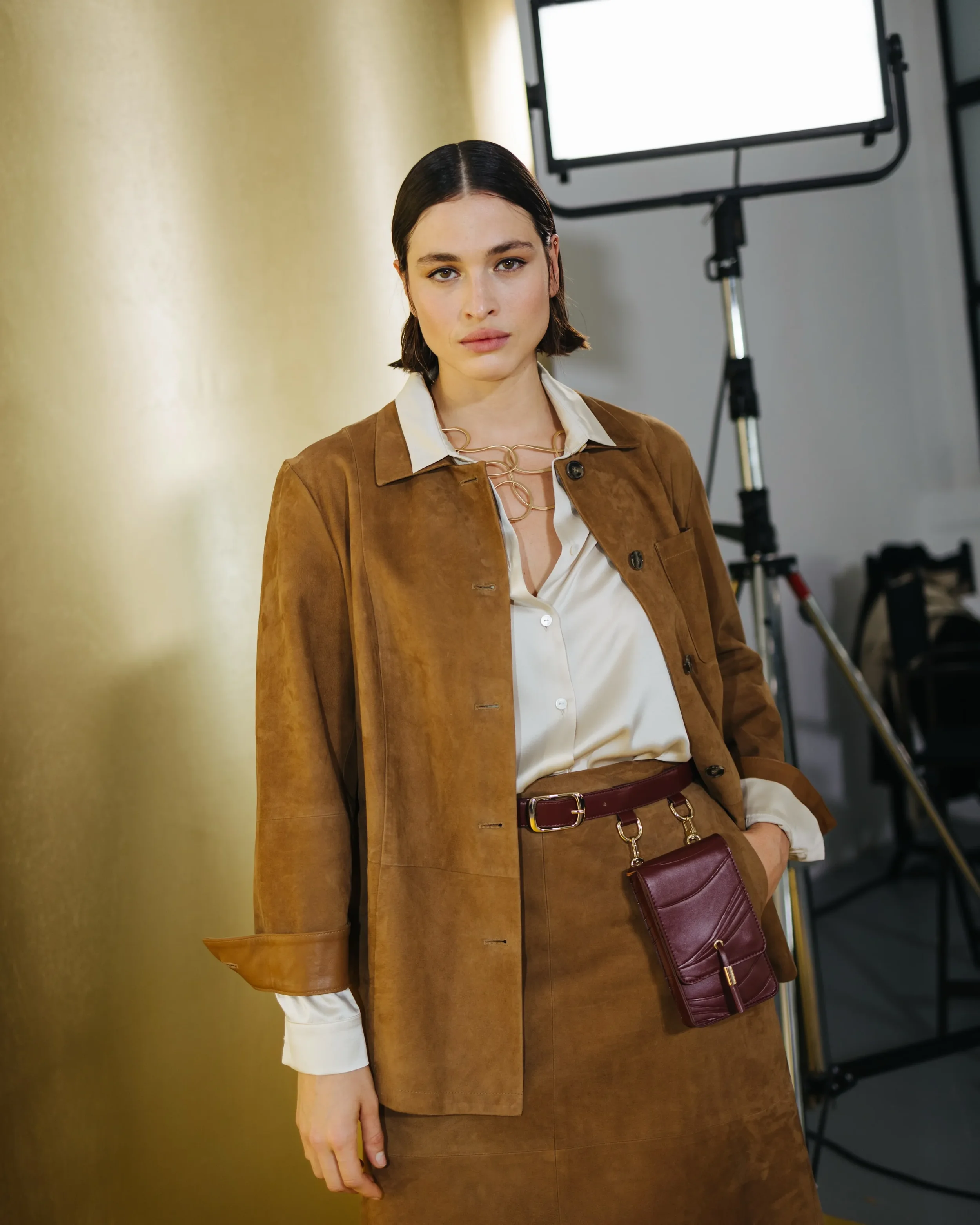 Scatto backstage con modella in giacca di camoscio marrone, camicia in seta e borsello da cintura in pelle, illuminata da faretti da studio davanti alla quinta dorata della sfilata Marina Rinaldi.