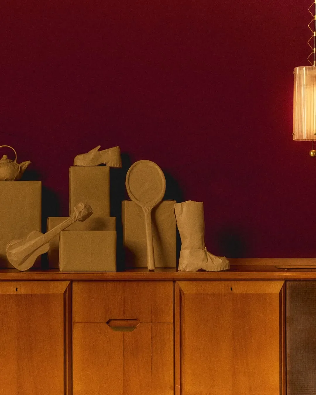 Set design e props styling per la campagna ADV Hogan 'The Wrapped Edit'. Oggetti quotidiani e sculture avvolti in carta avana disposti su una credenza in legno con sfondo borgogna.