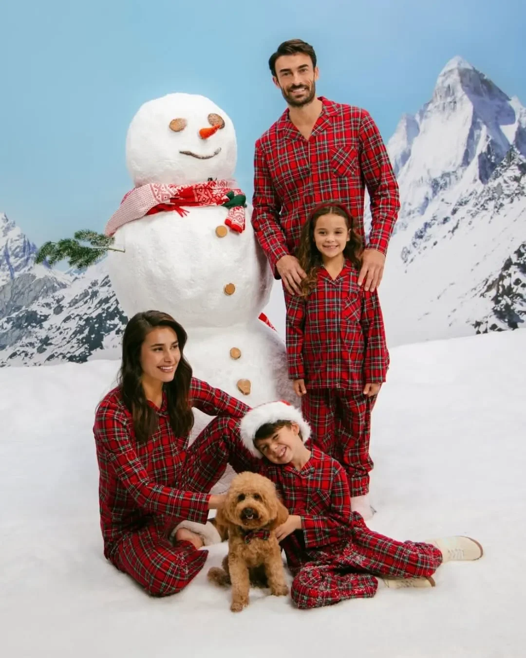 Famiglia in coordinato pigiama tartan rosso natalizio Yamamay in posa con un cagnolino e un pupazzo di neve su un set fotografico con paesaggio innevato.