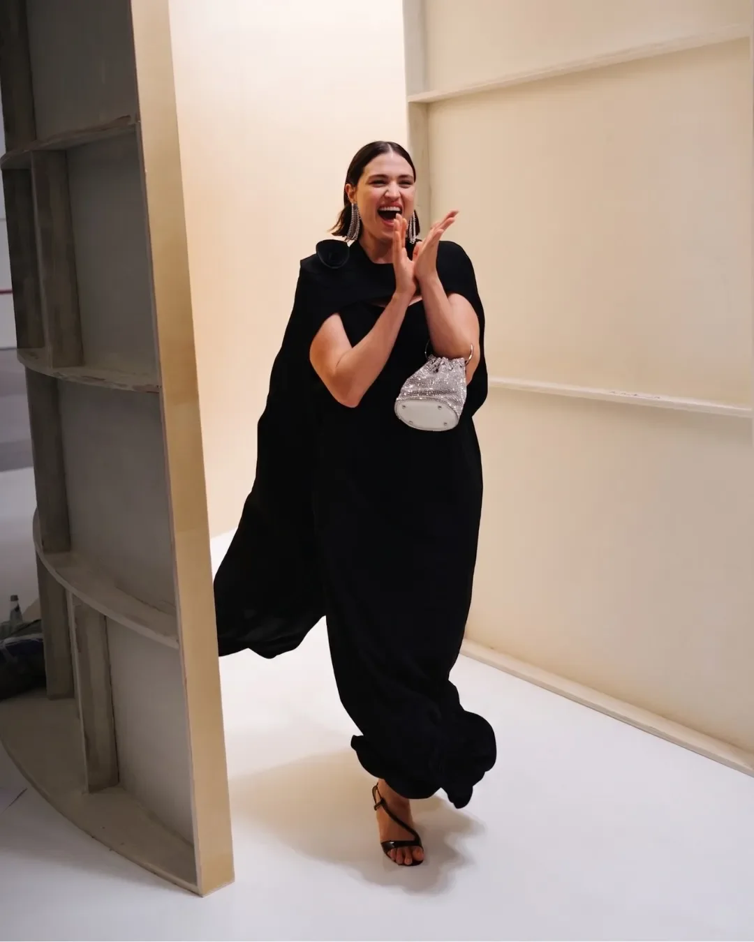 Modella sorridente ed entusiasta esce dalle quinte in legno del set design, indossando un abito nero con mantella e una borsetta gioiello in cristalli brillanti per la sfilata Marina Rinaldi