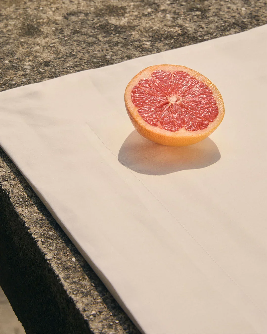 Composizione food e props styling con un mezzo pompelmo rosa adagiato su tessuto coordinato beige, fotografato su una superficie in cemento sotto la luce solare diretta.