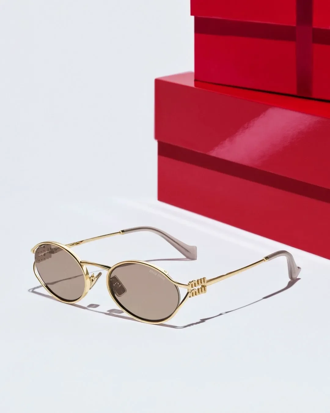 Fotografia still life di occhiali da sole ovali Miu Miu in metallo dorato con logo sull'asta, posizionati su superficie bianca davanti a una pila di scatole regalo rosse con nastro in raso per Sunglass Hut.