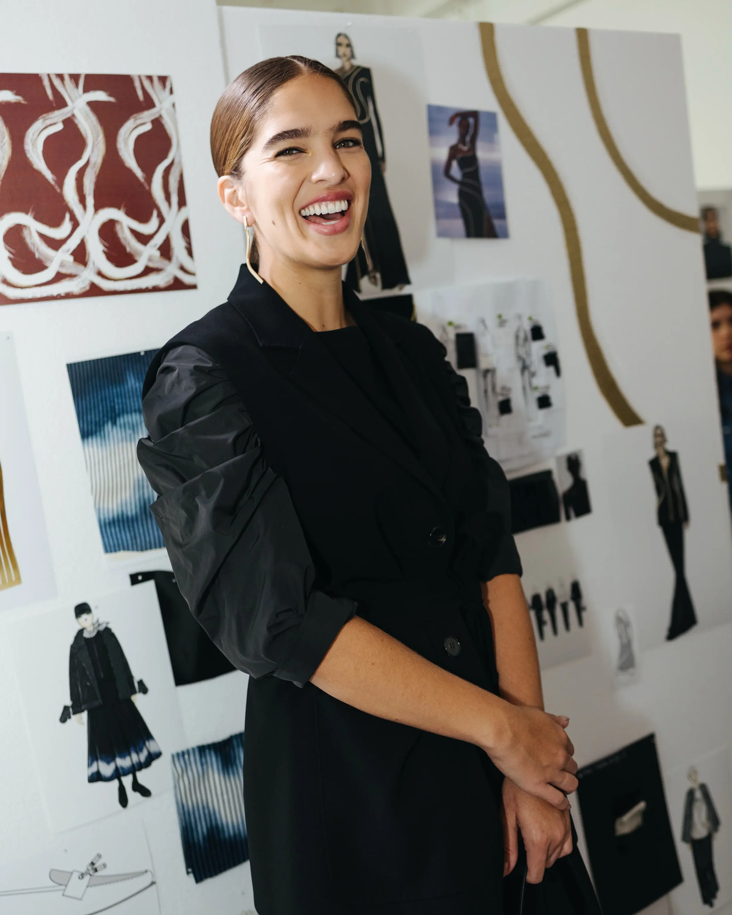 Modella nel backstage della sfilata Marina Rinaldi con gilet sartoriale nero e maniche a palloncino, posizionata davanti alla parete espositiva con bozzetti e studi di collezione.