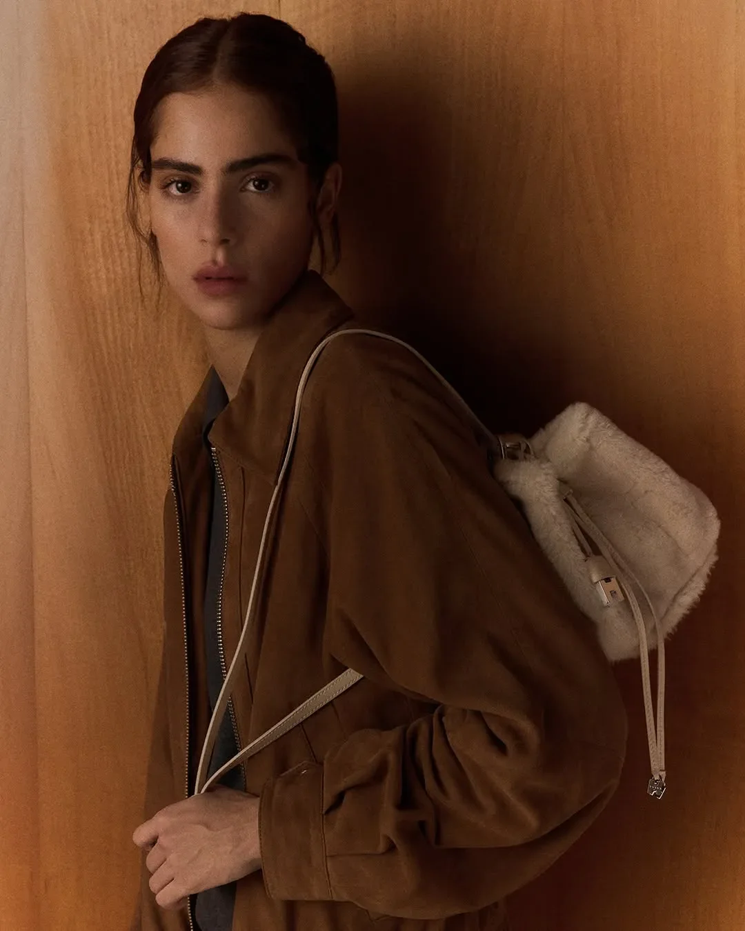 Scenografia d'interni e ritratto editoriale per la campagna ADV Hogan. Modella in giacca in camoscio marrone con zaino in pelliccia shearling beige su sfondo in legno.