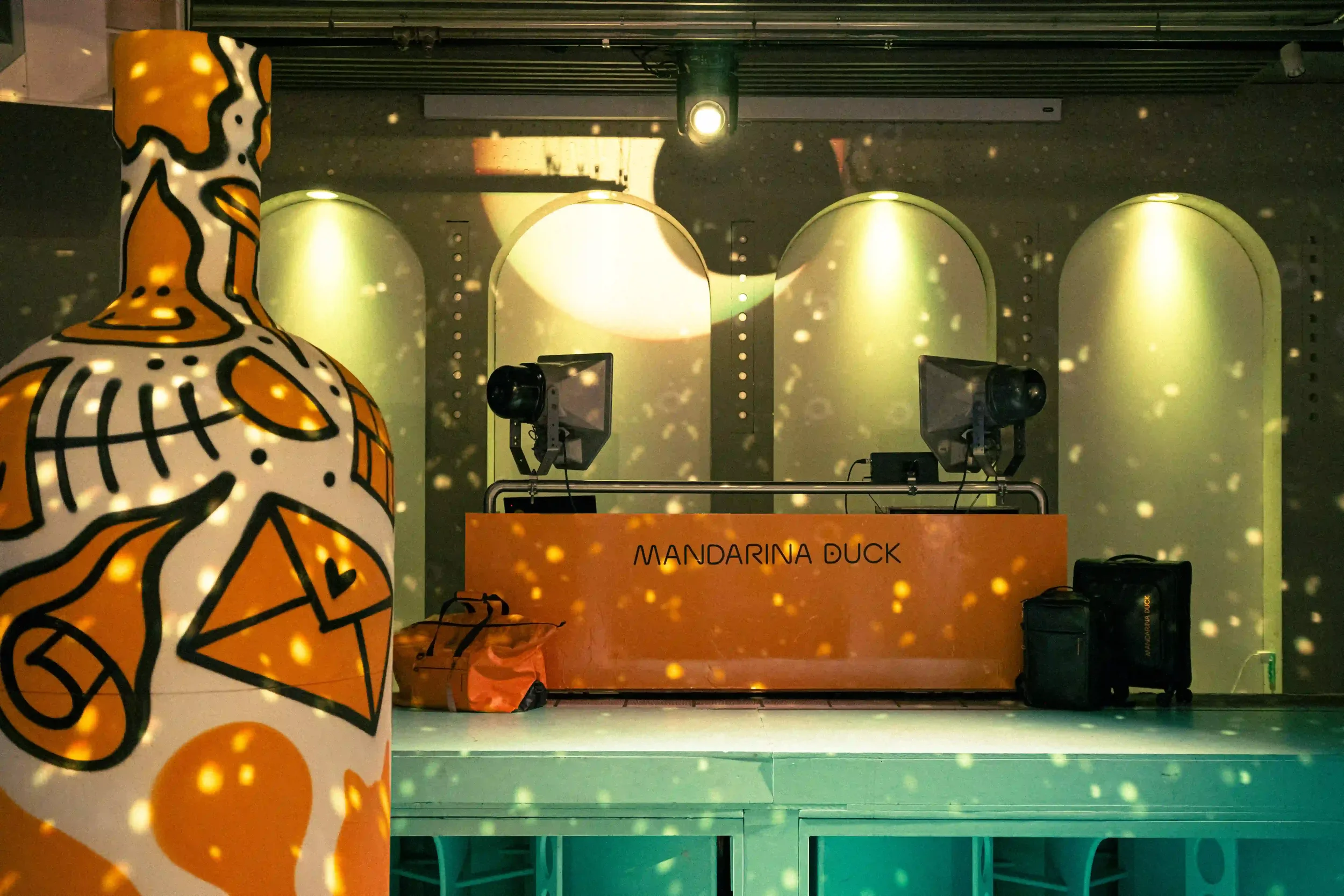 Set design della console DJ arancione Mandarina Duck allestita con valigie d'archivio, giochi di luce stroboscopici e scultura decorata a graffiti.