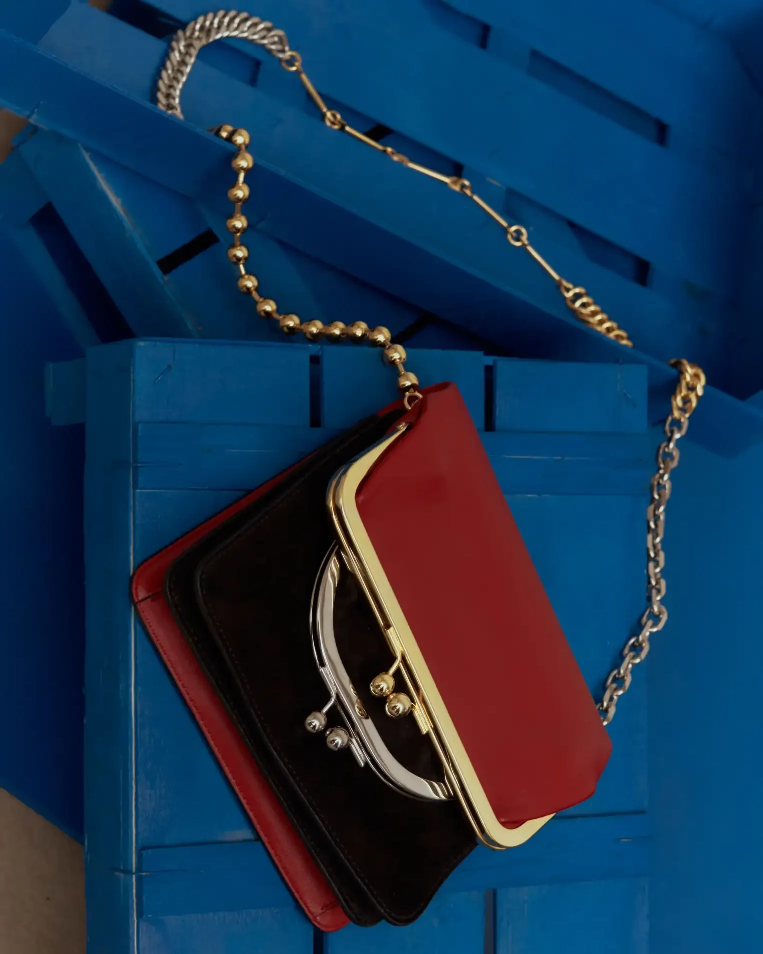 Fotografia still life di una borsa pochette bicolore in pelle rossa e camoscio marrone scuro con chiusura metallica vintage e tracolla a catena dorata, adagiata su cassette in legno dipinte di blu intenso. Set design editoriale per Le Mile Magazine.