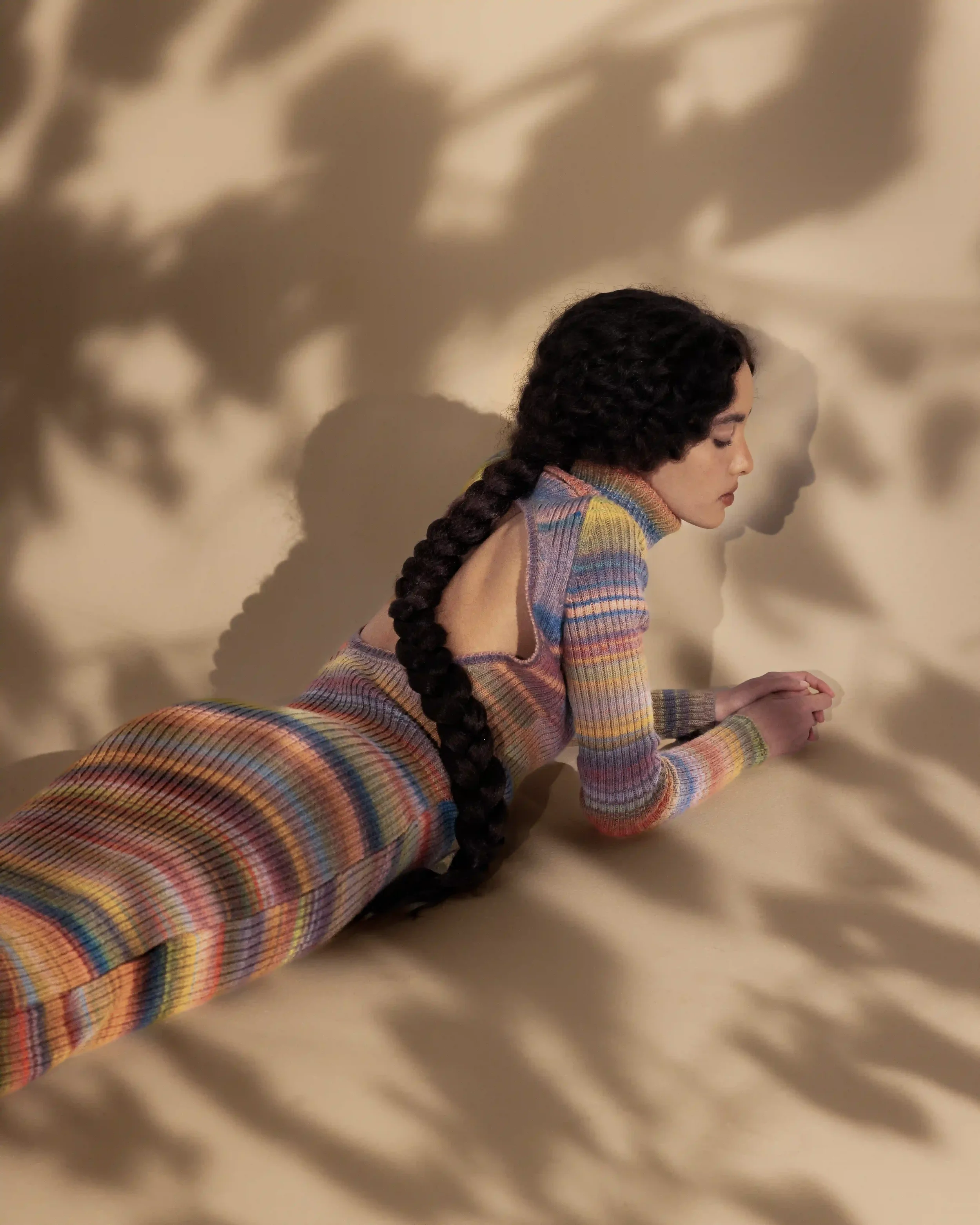 Scenografia e styling per editorial Laneus con giochi di luce e ombre foliate. Modella stesa a terra con abito lungo in maglia a righe multicolore con schiena scoperta.