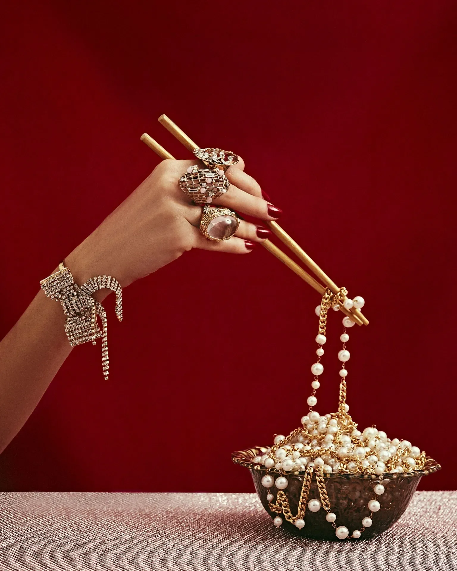 Scatto still life per la campagna Rosantica con mano impreziosita da anelli e bracciale che usa bacchette dorate per sollevare una collana di perle da una coppa.
