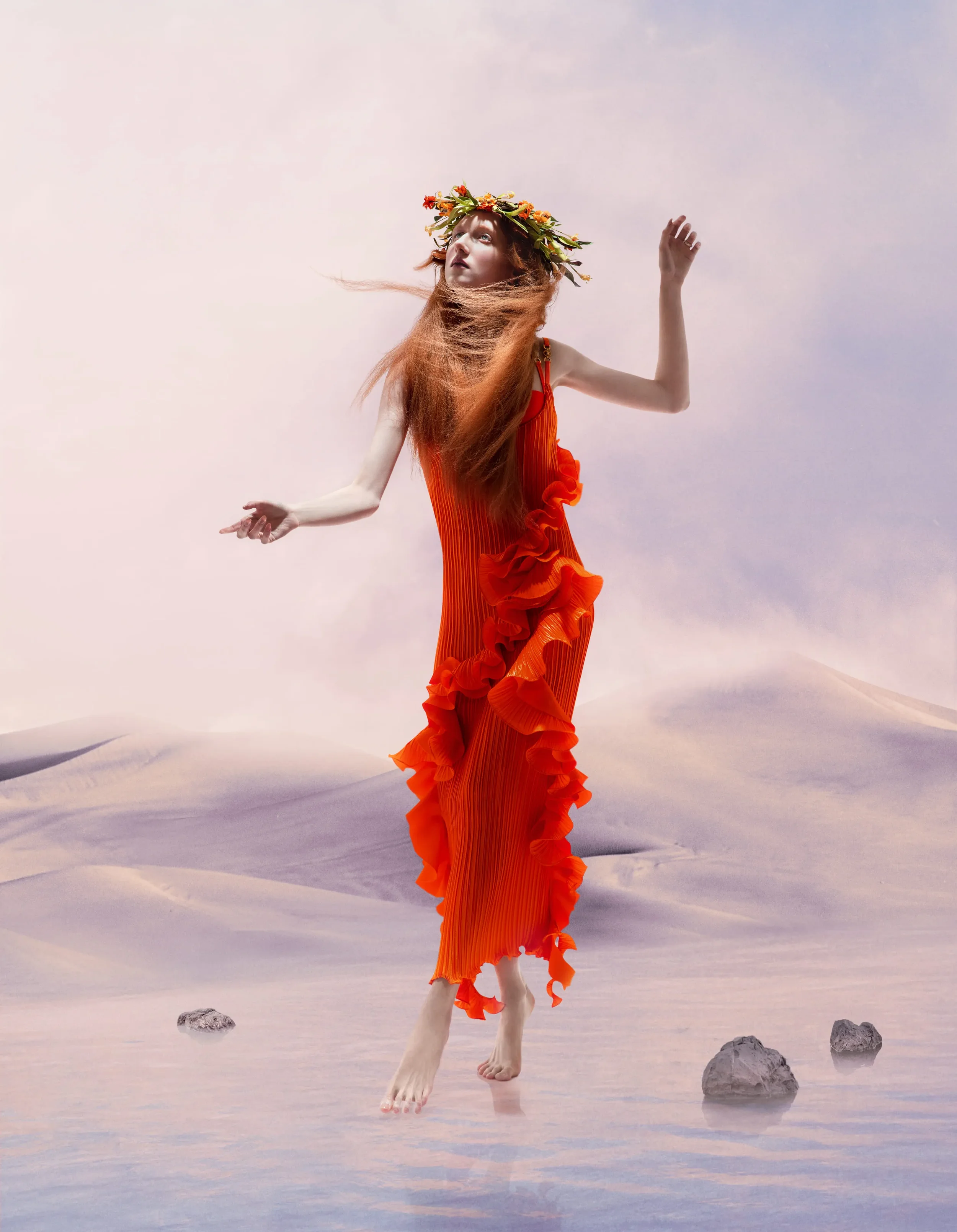 Fotografia di moda surrealista per Vogue Portugal con modella a piedi scalzi che indossa un abito plissé arancione con ruches e una corona di fiori, immersa in un paesaggio fiabesco di dune e acqua.
