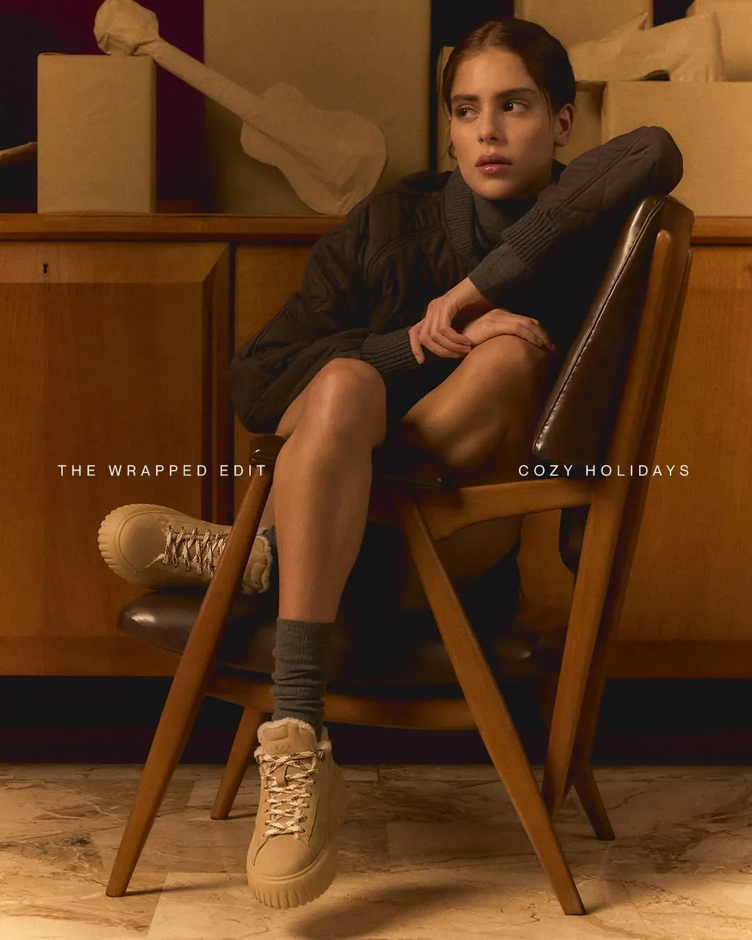Allestimento set per la campagna pubblicitaria Hogan 'Cozy Holidays'. Modella seduta su poltrona vintage in legno con sneakers beige e dettagli in shearling.