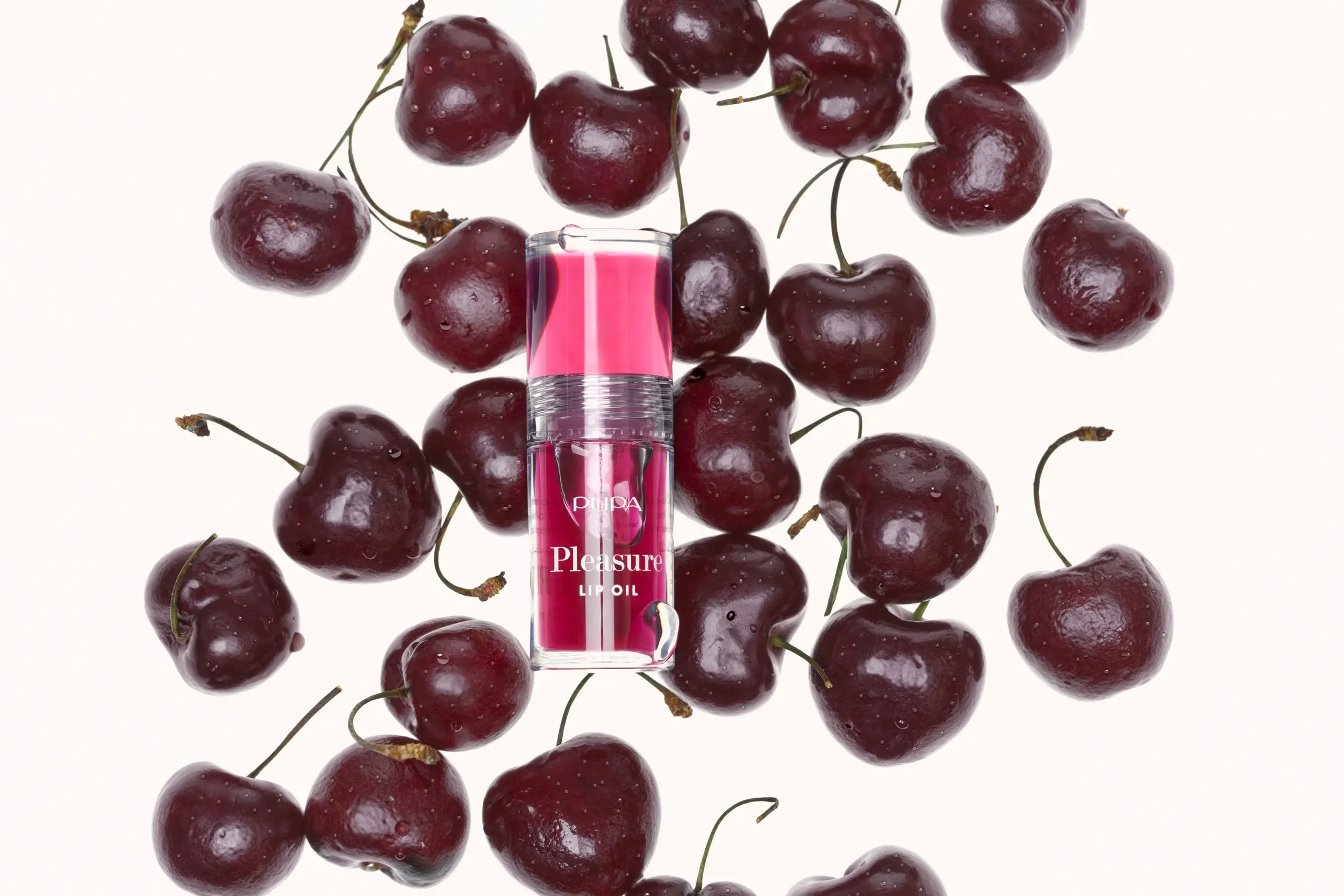 Fotografia still life con vista dall'alto del lucidalabbra Pupa Pleasure Lip Oil adagiato al centro di una composizione di ciliegie fresche su sfondo bianco luminoso.
