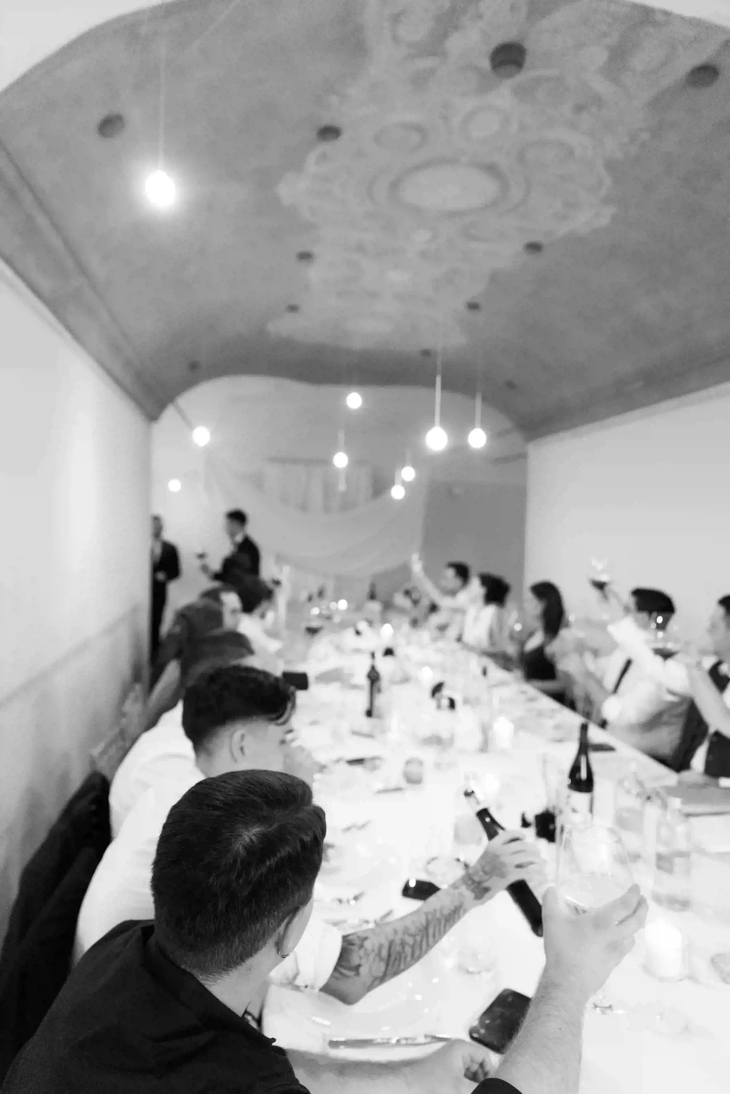 Scatto in bianco e nero d'atmosfera durante il brindisi degli ospiti seduti al tavolo imperiale nella sala ricevimenti della villa