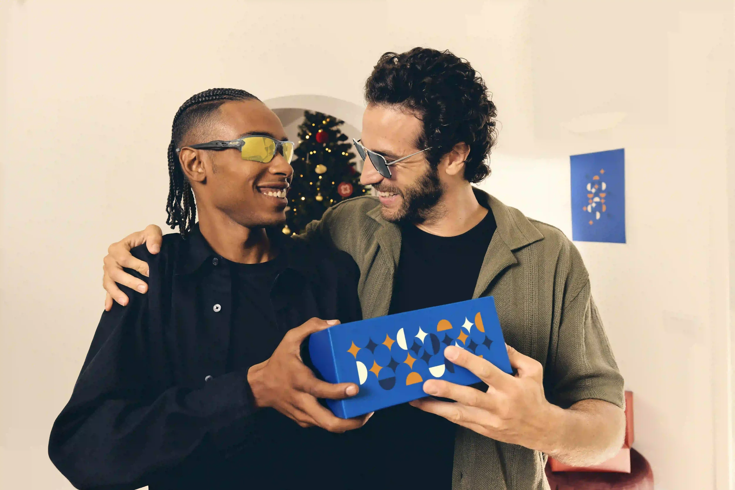 Scatto editoriale per la campagna natalizia di Sunglass Hut con due modelli che sorridono scambiandosi un pacco regalo blu di design, indossando occhiali da sole da uomo con lenti gialle e montatura metallica accanto all'albero di Natale