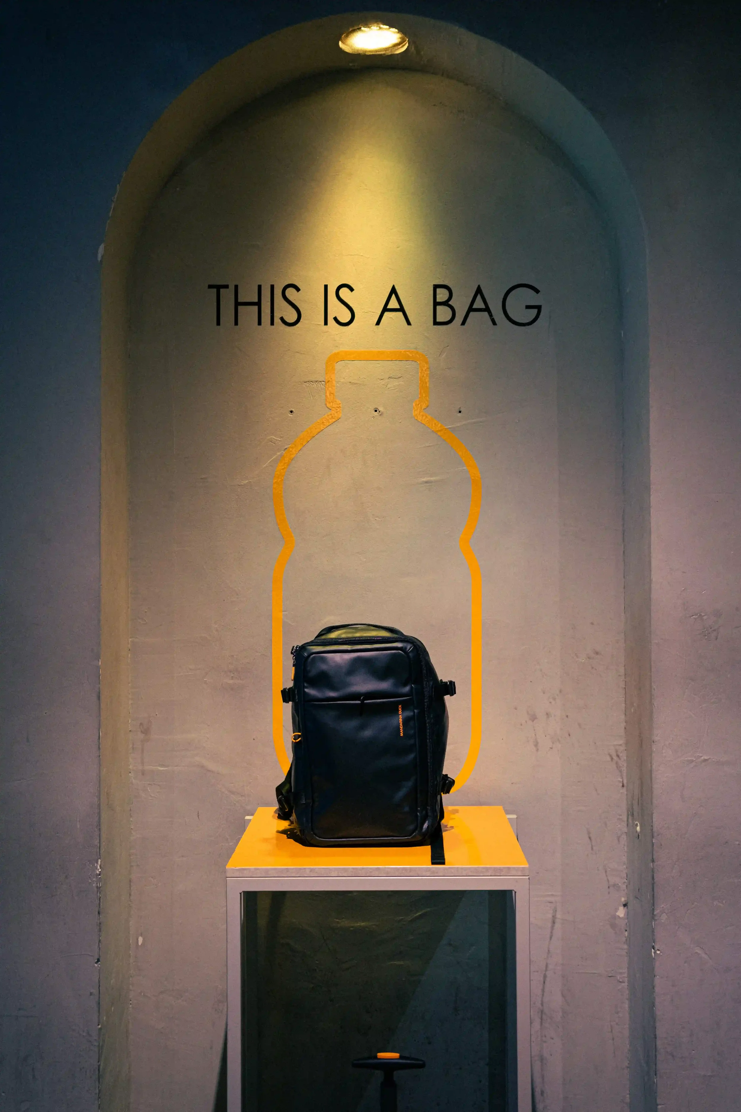 Display espositivo in nicchia ad arco per lo zaino EcoDuck sostenibile di Mandarina Duck a Berlino, con grafica a parete 'THIS IS A BAG' e sagoma di bottiglia riciclata.