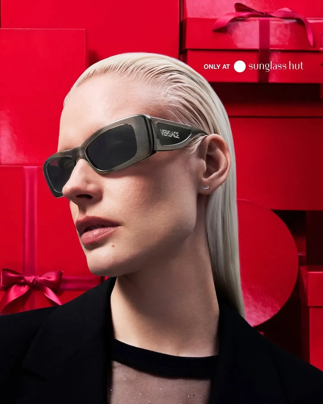 Primo piano di una modella bionda con capelli tirati indietro che indossa occhiali da sole Versace rettangolari grigio metallizzato, su sfondo di scatole regalo rosse per la campagna Sunglass Hut.