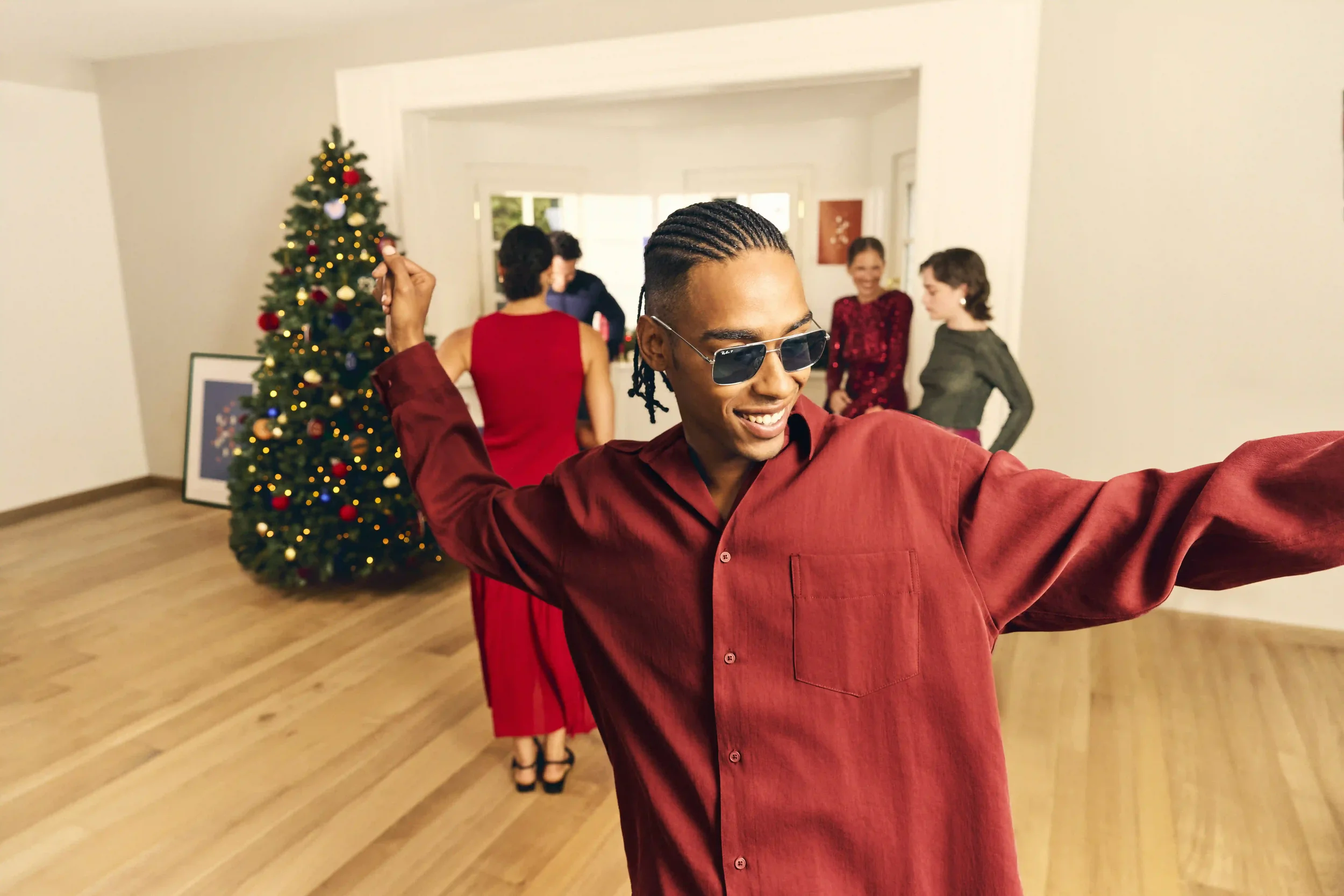Scatto dinamico di party per la campagna di Natale Sunglass Hut con modello che balla indossando occhiali da sole metallici rettangolari durante una festa tra amici.