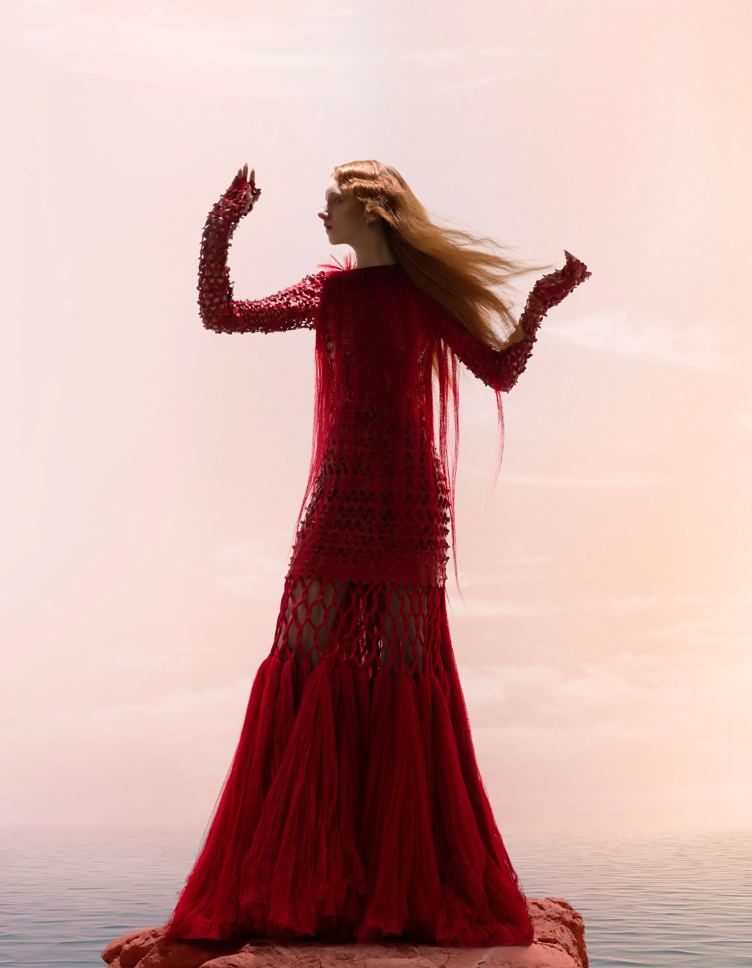Scatto moda editoriale per Vogue Portugal con una modella dai capelli rossi che indossa un lungo abito lavorato rosso con frange, in posa su una roccia in mezzo al mare al tramonto.