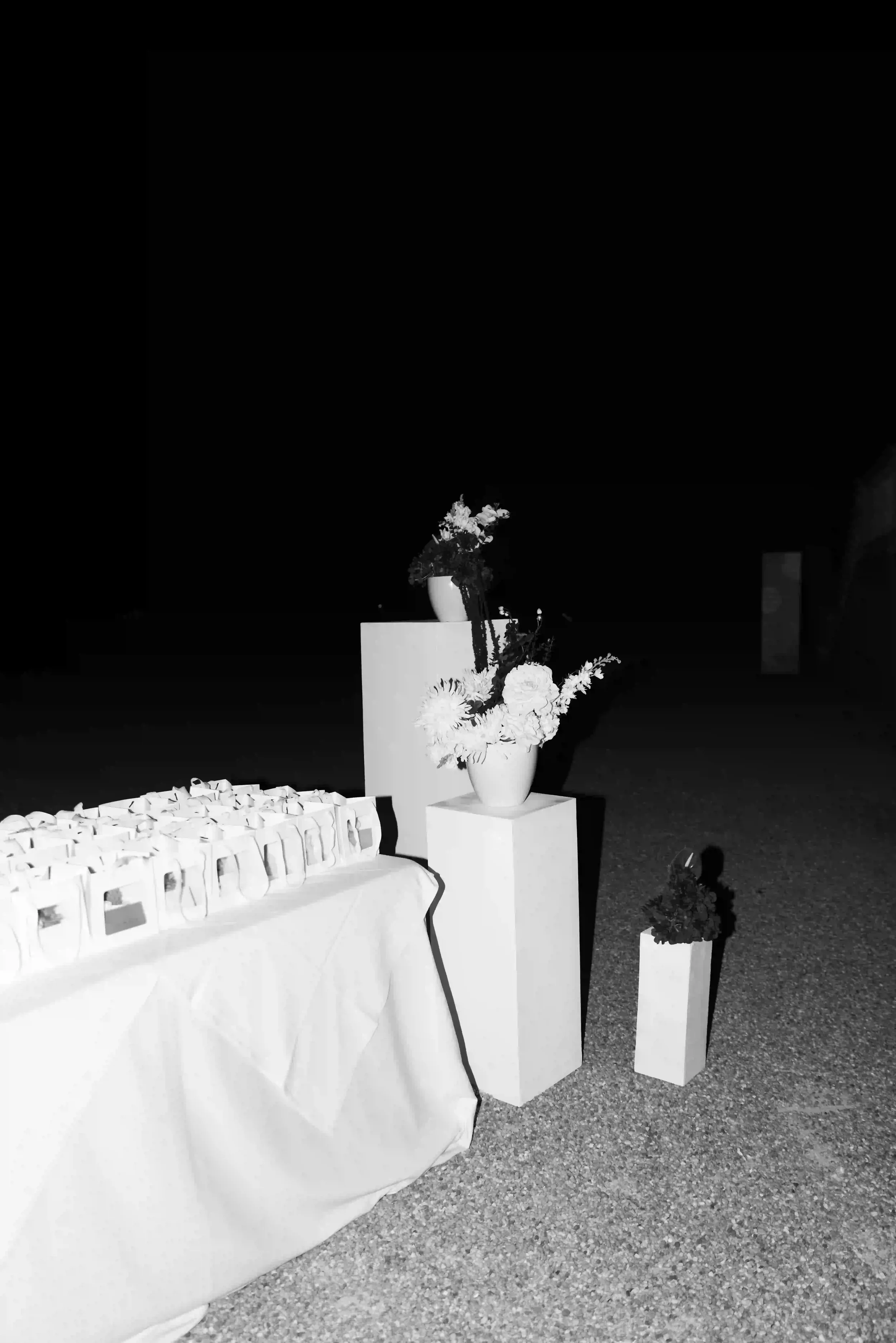 Fotografia in bianco e nero dell'allestimento scenografico notturno nel parco della villa, con il tavolo delle bomboniere affiancato da pedestali bianchi e composizioni floreali