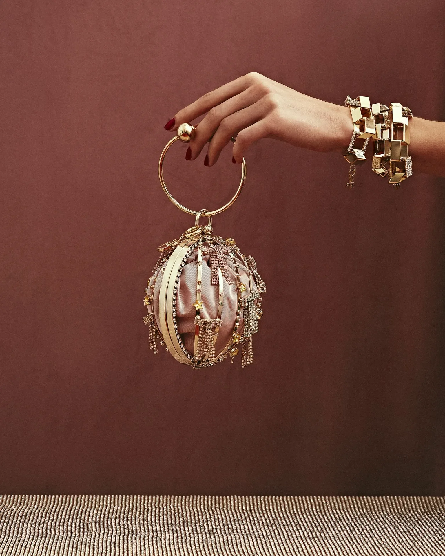 Scatto still life per la campagna Rosantica con mano adornata da bracciali dorati che tiene una borsa a sfera in metallo dorato con interno in raso rosa e pendenti di cristallo.