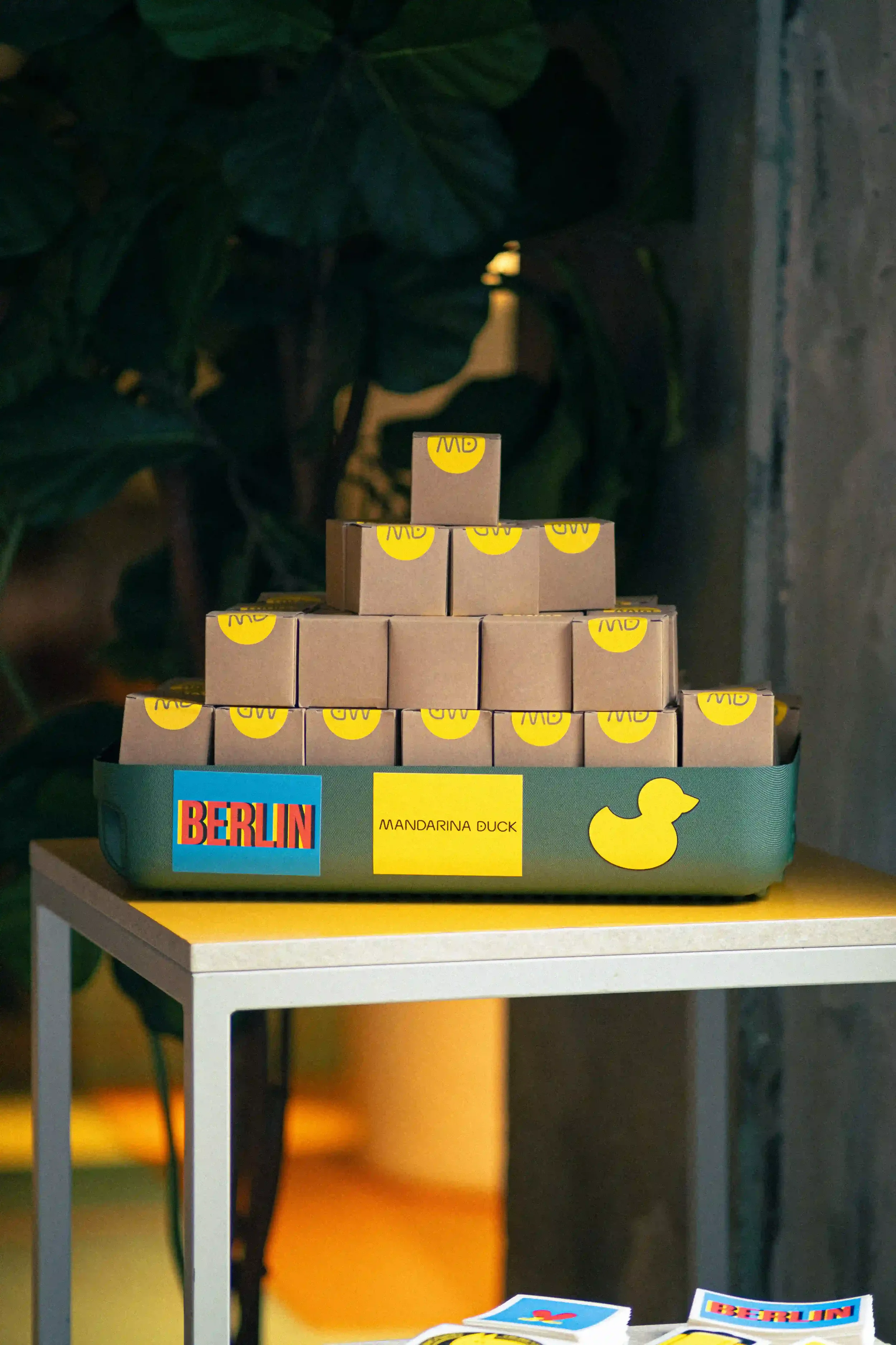 Composizione espositiva di scatole regalo gadget Mandarina Duck disposte a piramide su vassoio verde brandizzato 'Berlin' durante l'evento in Germania.