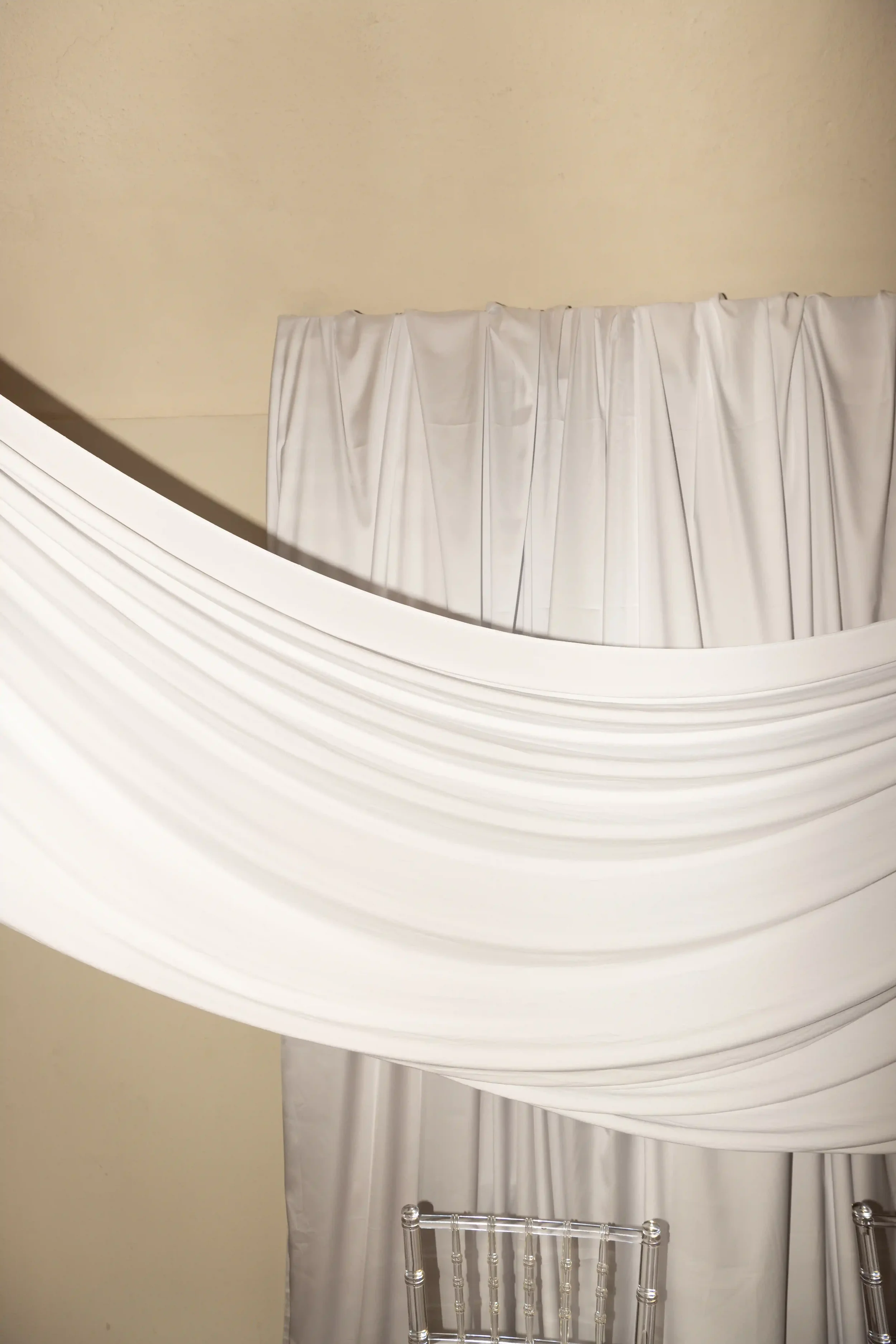 Particolare del set design interno con un ampio drappeggio scenografico in tessuto bianco satinato a parete e sedie Ghost trasparenti