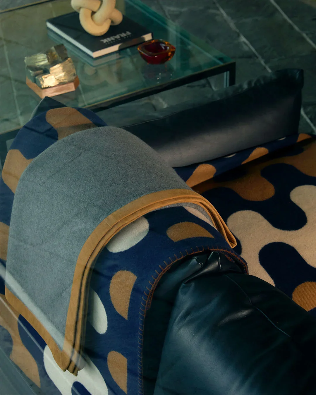 Set design d'interni e props styling con coperta plaid in lana a motivi geometrici blu e ocra drapata su un divano in pelle, vista dall'alto attraverso un tavolino in vetro con volumi e cristalli.